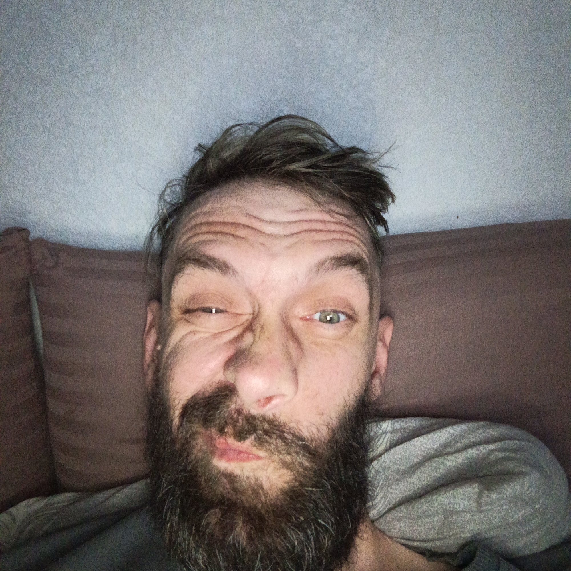 A selfie of a middle-aged man with a beard, lying on a bed or sofa. He has short, messy hair and is making a comical face with one eye squinted. The background is a light-colored wall and a brown pillow. There is no indication of this image being associated with the city of Марковский, Russia.