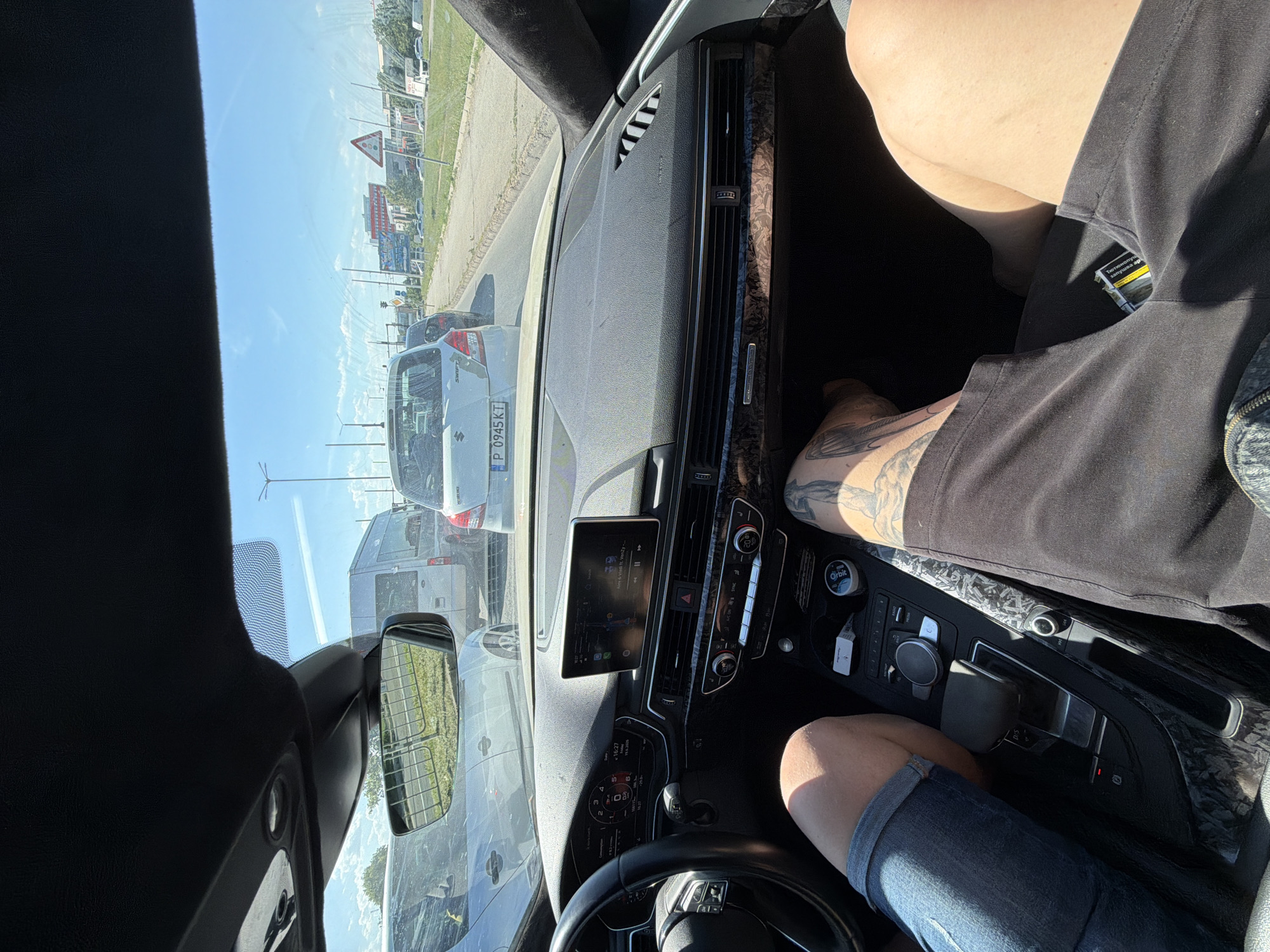The image is taken from inside a car, looking forward. The scene appears to be a sunny day, with a clear blue sky visible through the windshield. The car is in traffic, with other vehicles in front. The dashboard of the car is visible, featuring a central infotainment screen, climate controls, and a steering wheel. A person's legs and torso are visible in the driver's and passenger's seats. The person in the driver's seat has tattoos on their leg. A pack of cigarettes with text in Cyrillic script is visible in the pocket of a pair of black shorts. The surrounding environment suggests an urban or suburban setting, with buildings and road signs visible in the distance. The traffic light indicates the car is likely in a city or town. The license plate of a car in front is partially visible and reads "P 0945 KT". The overall mood appears relaxed, as suggested by the casual attire and the sunny weather. The location context provided is Dolno Ablanovo, Bulgaria.