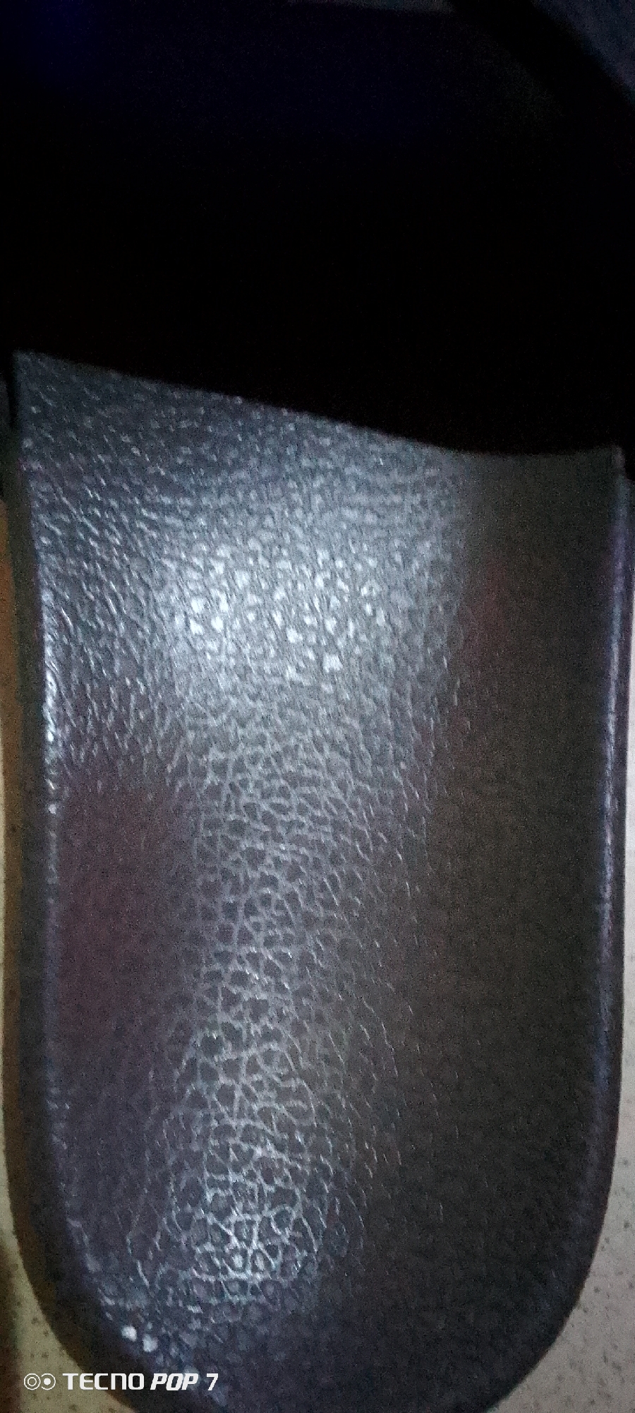 The image shows a close-up view of a dark, textured surface, possibly leather or a similar material. The texture is uneven, with a pattern of small, irregular indentations. A dark, out-of-focus area is visible at the top of the image. The bottom left corner shows a watermark that reads "TECNO POP 7". The location is listed as Unknown, Unknown.