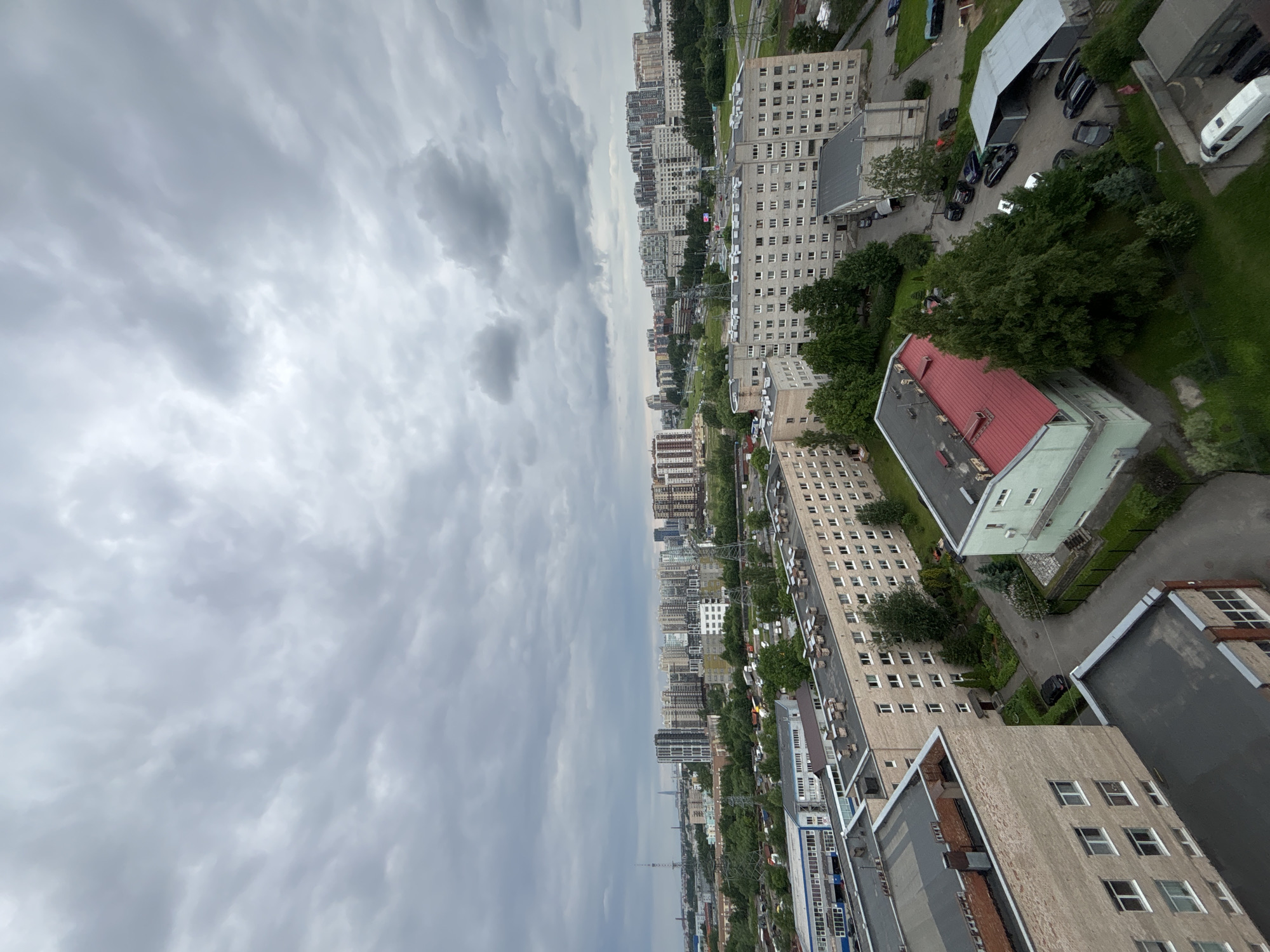 The image shows an aerial view of a city under a cloudy sky. In the foreground, there are several apartment buildings with flat roofs. Some buildings are light beige, others are darker brick. One building in the middle features a bright red roof. Beyond these buildings, there is a dense green area with trees. In the background, a large expanse of apartment blocks stretches across the horizon, with some taller modern buildings interspersed. There are also signs of industrial or infrastructure elements like power lines and towers visible in the distance. Several cars are parked in the courtyards and on pathways between the buildings. The overall scene depicts a residential area within a city.