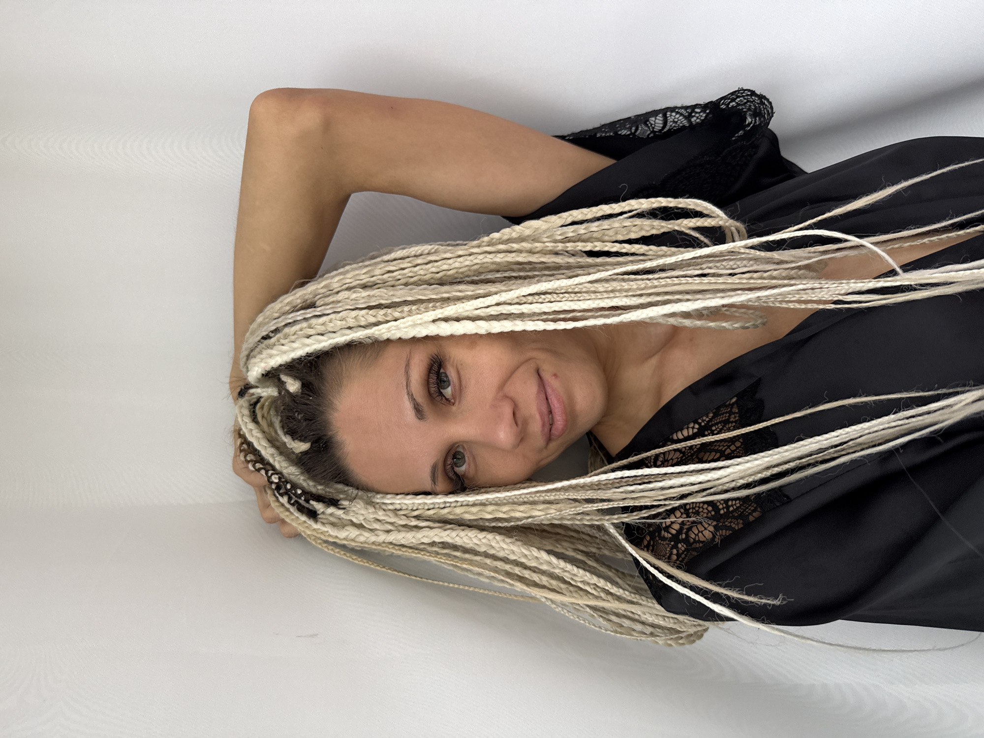 A medium shot captured from a low angle shows a woman with blonde box braids standing in the corner of a room. She is wearing a black robe with lace detailing on the neckline and sleeves. Her right arm is raised and bent, with her hand resting on the back of her head. The woman is looking towards the camera with a slight smile. The background is a plain white wall, with the corner of the room clearly visible. No specific location in Yaroslavl, Russia is discernible from the image.