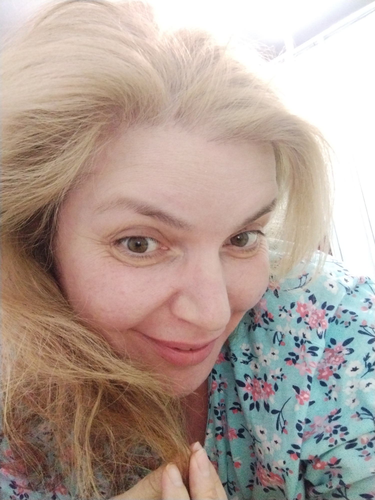 A close-up, waist-up selfie of a fair-skinned woman with blonde hair and light brown eyes, is shown. She is wearing a light blue floral-patterned shirt with pink and white flowers. Her head is tilted and she is looking towards the camera. The background is out of focus and appears to be a room. The image is well-lit, with the light source seemingly coming from the top left. The woman's expression is neutral, and her mouth is slightly open, revealing her teeth. The overall scene is casual and personal.