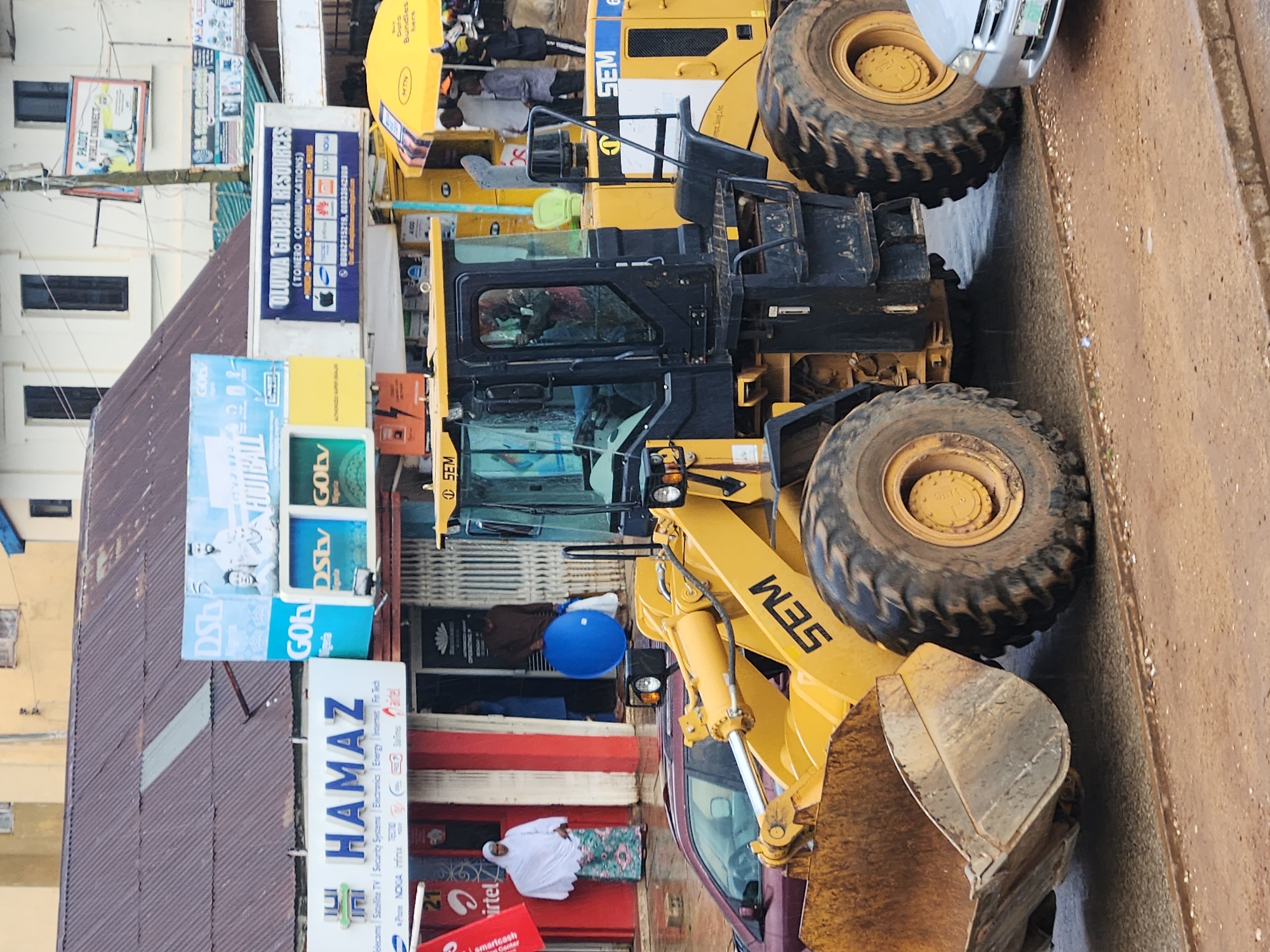 The image depicts a street scene in Jos, Nigeria, dominated by a large yellow SEM loader parked on a road. The loader's bucket is lowered to the ground, and its tires are large and rugged. In the background, various shops and businesses line the street, with awnings and signage visible. One prominent sign reads "HAMAZ," advertising telecommunications and electronics. Another sign, "OLUWA GLOBAL RESOURCES," also advertises communications services. Several people are seen in the background, some walking, and one woman in a white hijab and patterned skirt stands near a red shop front. A blue plastic sphere is being carried by a person entering a shop. A silver car is parked to the right of the loader. The environment appears to be overcast or rainy, as suggested by the wet road surface and the presence of a yellow umbrella in the background.