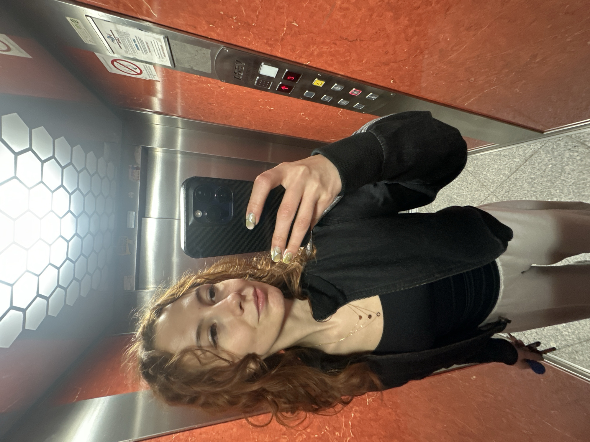A selfie photograph depicts a woman in an elevator. The elevator's interior features stainless steel walls with a honeycomb pattern on one side and a control panel on the other side. The control panel is set against a red, mottled surface. The woman is holding a black smartphone in her left hand to take the picture. She has reddish-brown, curly hair and is wearing a black jacket and a black top. A delicate gold necklace is visible around her neck. She is facing the camera. Part of her lower body, including grey pants, is visible in the reflection of the elevator walls. It is probable the elevator is located in Novi Sad, Serbia but is impossible to confirm based on the photograph.