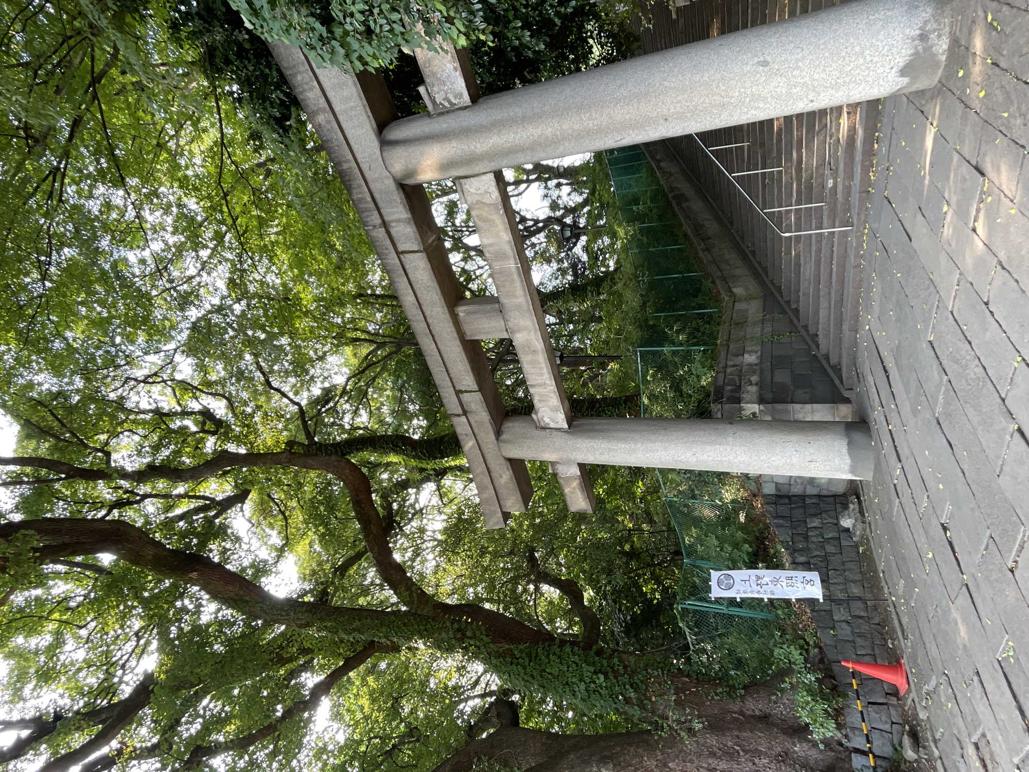 In Taito, Japan, a stone torii gate stands at the base of a wide stone staircase leading upwards. The torii gate is a traditional Japanese structure, with two upright posts supporting a lintel. The staircase is flanked by stone walls and a metal fence, with lush green trees and foliage overhead, filtering the sunlight. A white banner with Japanese characters hangs from the fence on the left side of the image, and a bright orange traffic cone is placed on the stone pathway in front of it. The pathway is made of large, rectangular stone slabs, with a few fallen leaves scattered across them. The scene appears to be a serene and historical location, possibly a temple or shrine entrance.
