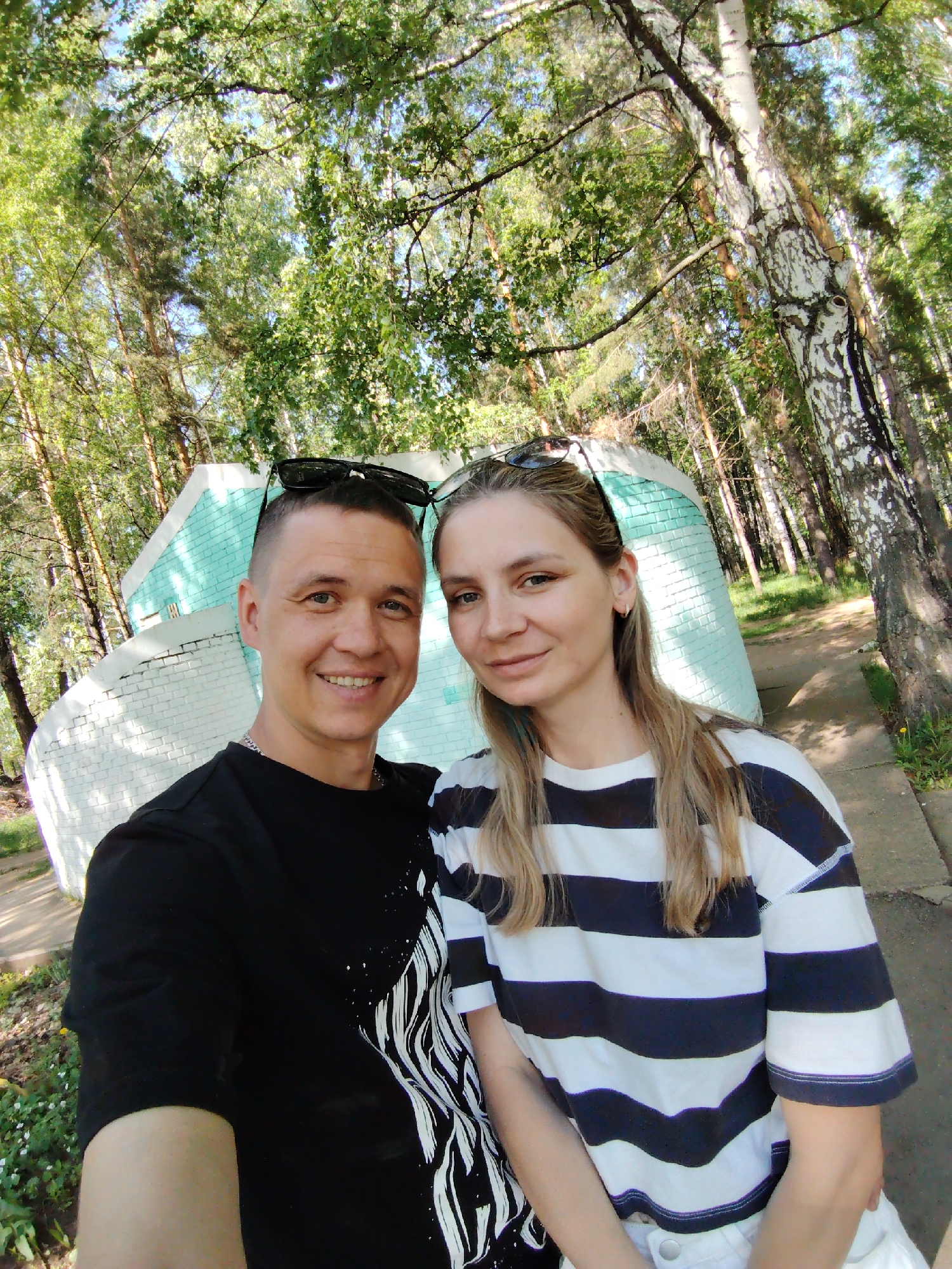 A selfie shows a fair-skinned male and female, with sunglasses perched on their heads, posed in front of a small, light blue and white brick building. The male is wearing a black t-shirt with a white design and a silver chain. The female is wearing a horizontally striped navy blue and white shirt. They are standing on a concrete walkway amidst trees, with the trees filling the background. The picture was taken in the city of Chaikovsky, Russia.