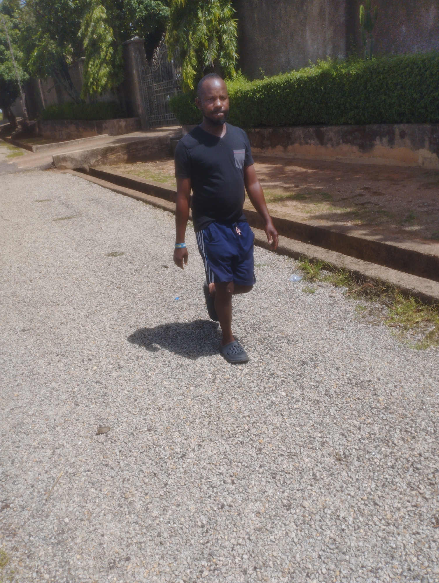 A man is walking on a gravel path in front of a stone wall in Jos, Nigeria. He is wearing a black t-shirt and blue shorts. He has a blue bracelet on his left wrist and dark sandals on his feet. There is a shadow of the man on the ground behind him. The background shows trees, a gate, and a hedge. The path appears to be a road or driveway. The lighting suggests it is daytime with the sun high in the sky.