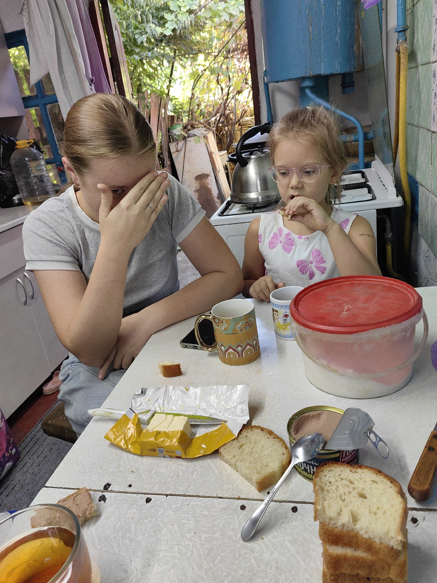 A young woman and a child are seated at a table, seemingly in a rustic outdoor kitchen area. The woman, with her arm covering her face, wears a grey t-shirt and blue shorts. The child, wearing a white top with pink bow details, has glasses and is eating something. On the table are several items including slices of bread, a can of pâté with a spoon, butter, mugs, and a large red-lidded container. The background shows a kitchen with a stove, a kettle, and shelves, suggesting a somewhat aged or rural setting. The overall environment appears to be outdoors or in a poorly maintained indoor space, possibly a dacha or an older residential building. The specific location in Shymkent, Kazakhstan is not discernible from the visual cues.