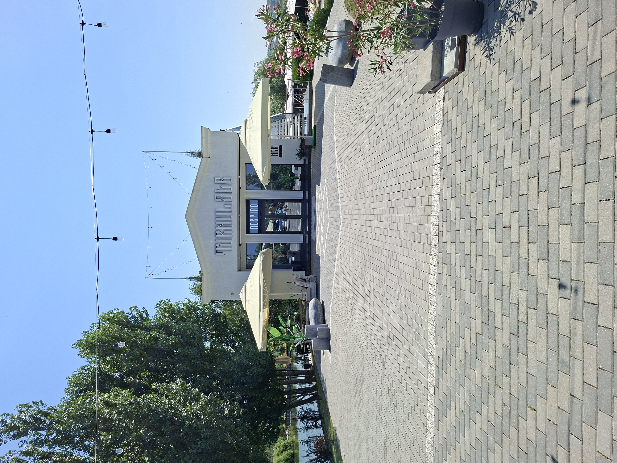 The image shows an outdoor scene in Costești, Moldova, on a clear, sunny day. In the foreground, a paved walkway made of grey and tan rectangular bricks leads towards a white building. The building features large glass doors with the word "RESPIRO" above them and "TURNUL BAR" in large illuminated letters on its facade. Two large, cream-colored umbrellas are positioned in front of the building. To the left of the building, a body of water, likely a river, is visible bordered by trees. To the right, there are potted plants with pink flowers and more outdoor seating with umbrellas. String lights are visible suspended across the scene. The overall impression is of a pleasant, modern outdoor establishment, possibly a bar or restaurant, situated near a waterfront. There are no visible people engaged in any specific actions.