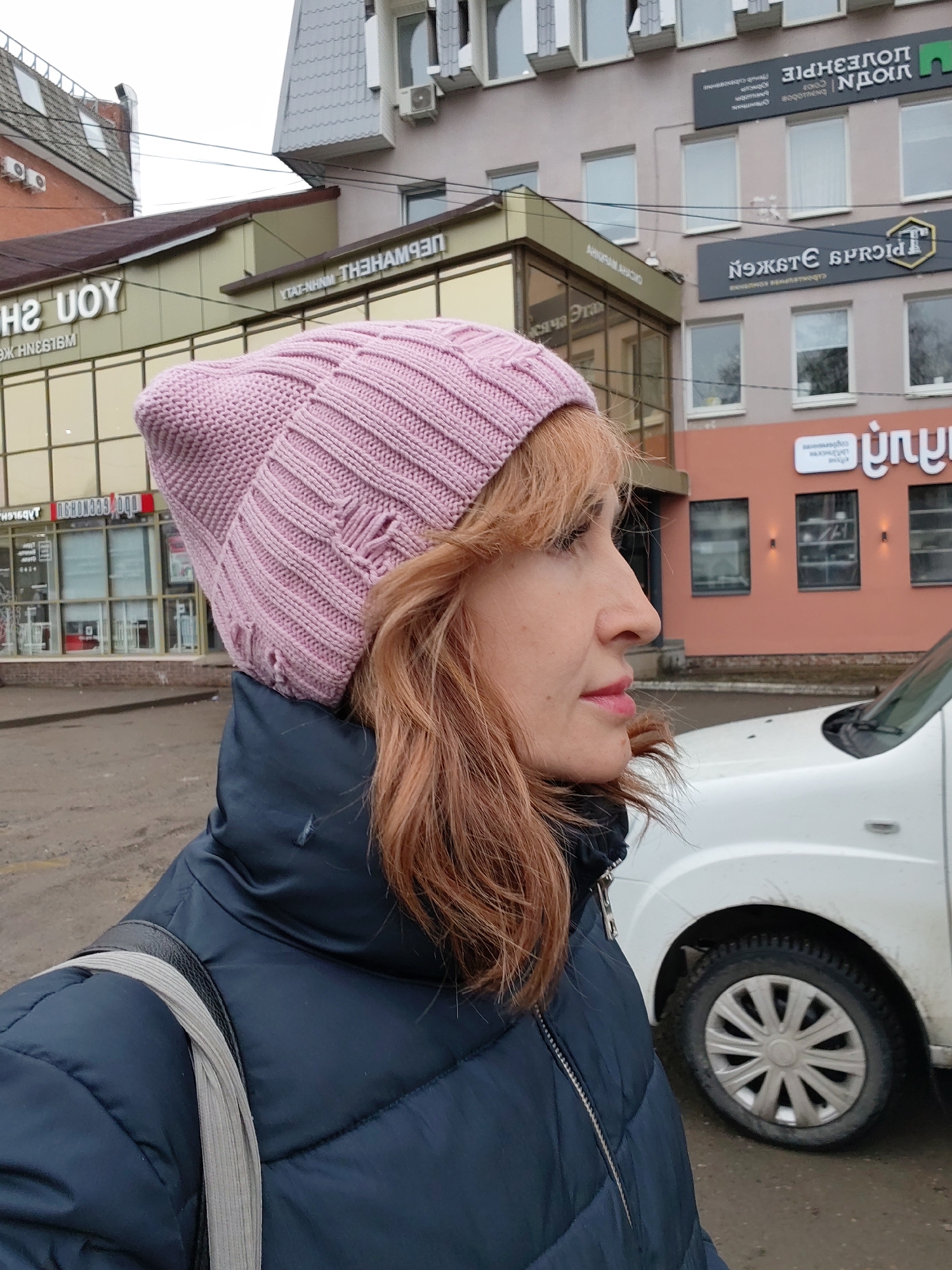 The image depicts a woman in profile, wearing a pink knitted hat and a dark blue padded jacket. A gray strap, presumably from a bag or purse, is visible on her shoulder. She is outside, near a street, with a white car partially visible to the right of the frame. The backdrop consists of buildings with signage in a Cyrillic alphabet.
Based on the signage visible, the image is in Chaikovsky, Russia. Specifically, signs like "HUOY," "YTAT-NHNM THEHAМАЗП," "AHIRAN AHACIO," "ЦОУЕЗНΡΙΕ ПОУЕЗНΡΙΕ," "NOMSTE SPROIN," and "диз" are identifiable in the urban environment.