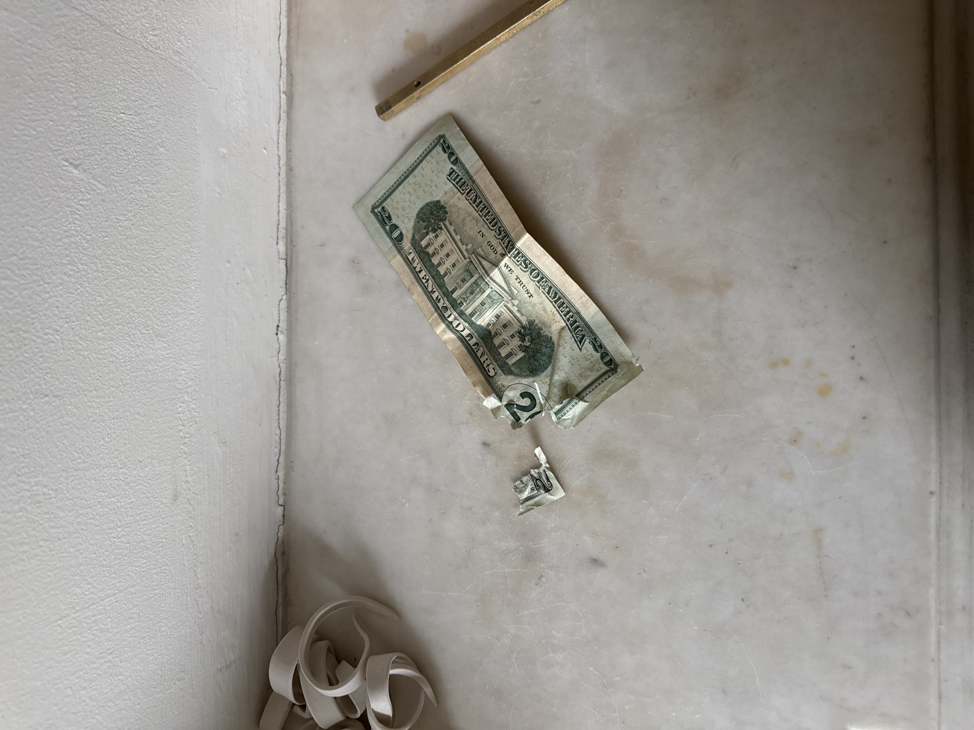The image captures a damaged twenty-dollar bill lying on a light-colored, possibly marble or stone, surface. The banknote is torn across the middle, with a small, detached fragment showing the number "20" positioned just below the larger piece. Visible text on the main section of the bill includes "THE UNITED STATES OF AMERICA", "IN GOD WE TRUST", and "TWENTY DOLLARS".
Above the torn currency, a golden, rectangular object, resembling a metal bar or a part of a tool, is resting on the same surface. In the lower-left corner of the frame, several white, curled strips of material, possibly paper or plastic shavings, are clustered together. The background on the left features a textured, light-colored wall, which meets another surface or wall at a discernible corner line. The overall setting appears to be an indoor space, possibly a utility area or workshop, given the nature of the items and surfaces. There is no explicit activity taking place, but the torn bill and waste material suggest a past event involving damage or discarding. The lighting is bright and even.