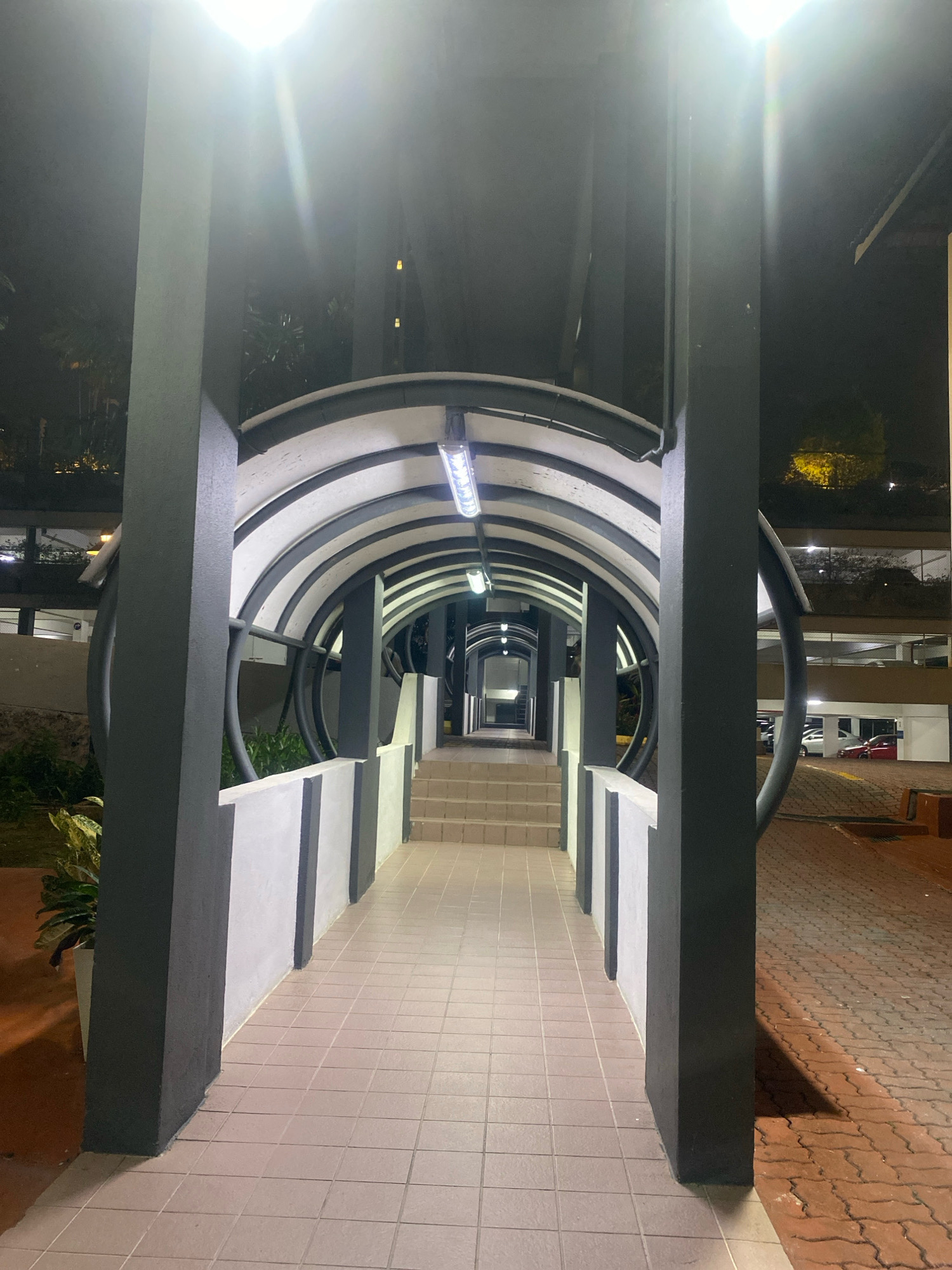 The image shows a covered walkway or bridge at night, illuminated by overhead lights. The walkway has a curved, tunnel-like structure with a white roof and black supports. The floor of the walkway is tiled with square, light-colored tiles. To the right of the walkway, there is a paved area with interlocking red bricks, and beyond that, a multi-level parking garage is visible, with cars parked on its levels. Trees and foliage are present on the left side of the walkway, with some greenery visible through the open sides of the covered structure. The scene appears to be located in an urban environment, possibly an area with pedestrian access between buildings or to a parking facility. The bright overhead lights create a stark contrast with the dark surroundings and cast shadows.
