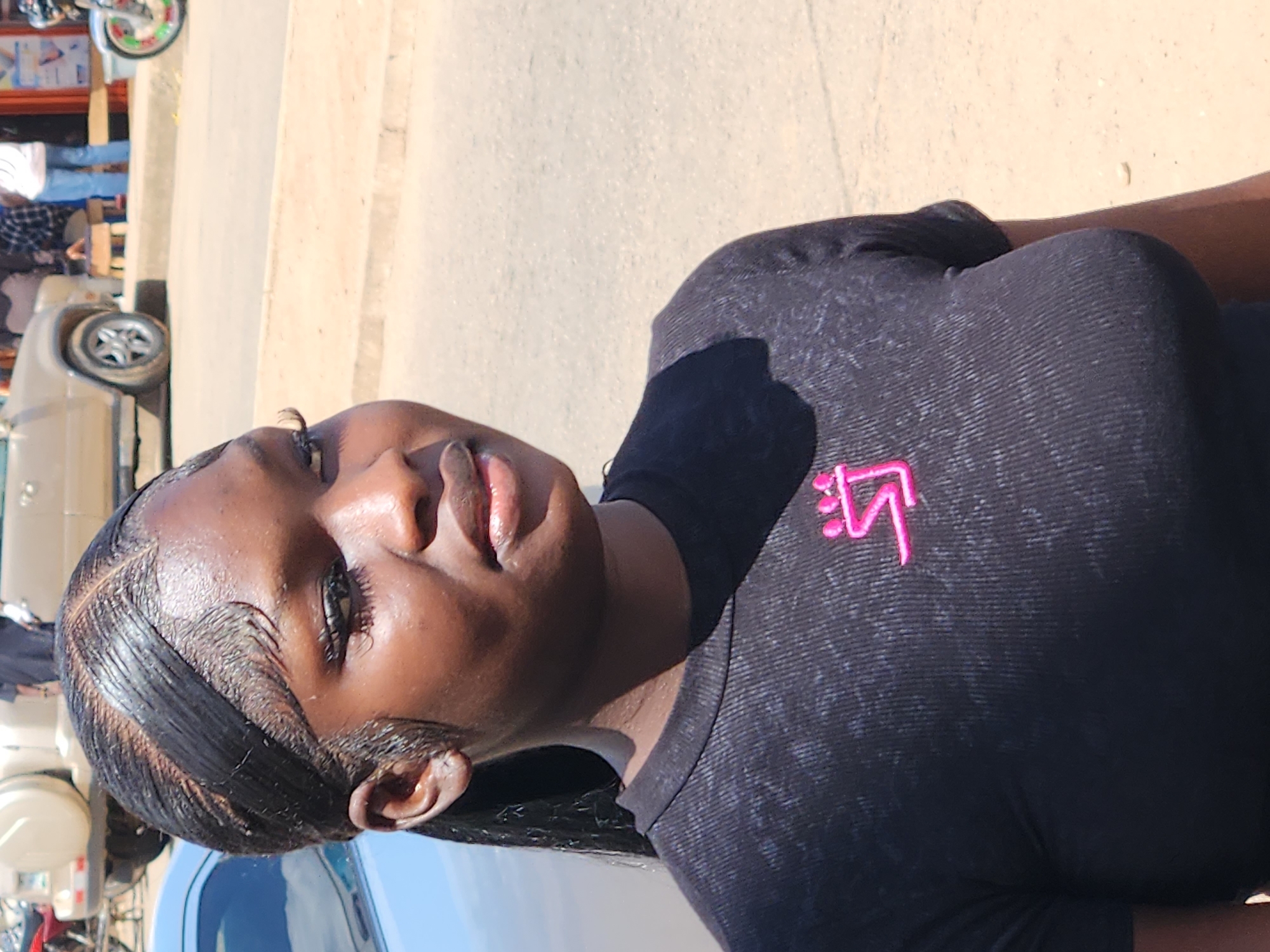 A medium close-up shot shows a young Black woman with her head turned to the right and slightly upwards, looking off-camera. She has dark, slicked-back hair with gelled edges and her eyelashes are prominent. She is wearing a black, ribbed t-shirt with a small, bright pink logo on the left side of her chest. The background is blurred, showing a street scene with a light-colored car, a motorcycle, and people walking in the distance. The lighting suggests it is daytime with bright sunlight casting a shadow from the woman onto the ground behind her. The environment appears to be an outdoor urban setting.