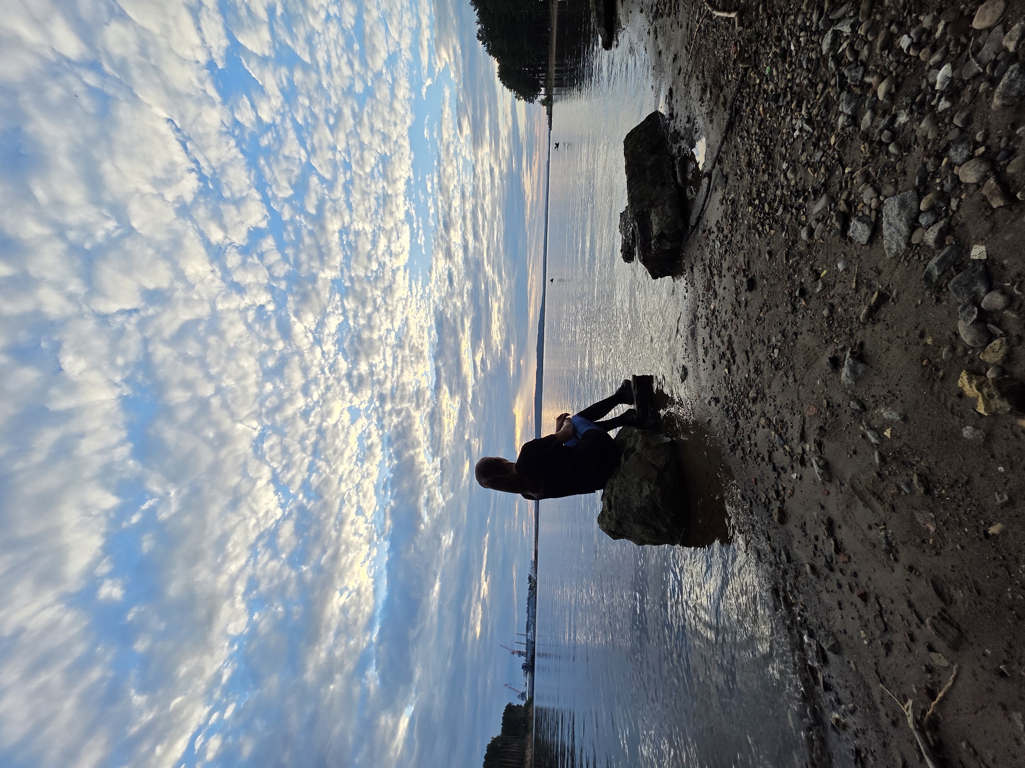 A person is sitting on a rock at the edge of a body of water, facing away from the camera. They are silhouetted against a sky filled with scattered white clouds and a hint of sunset. The water is calm, reflecting the sky. The shoreline is rocky and sandy. In the distance, across the water, there are trees on one side and industrial structures, including cranes, on the other. The overall scene evokes a sense of quiet contemplation. This appears to be the Kama River in Chaikovsky, Russia, near its industrial port.