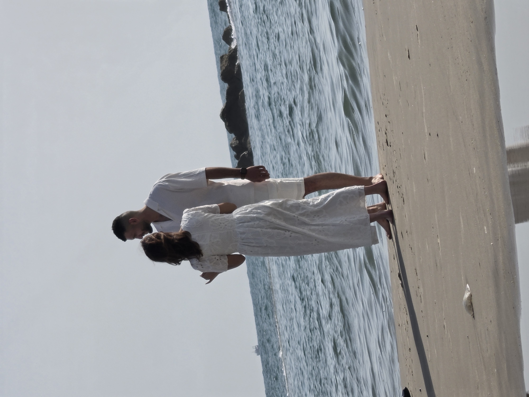 This image, when viewed in its correct orientation (rotated 90 degrees counter-clockwise), depicts a tender moment between a man and a woman on a beach or pier, likely in Bat Yam, Israel.
The two subjects are positioned close to each other, their faces angled towards one another, suggesting they are about to kiss or have just shared an intimate embrace. Both are dressed in all-white attire. The man, on the left, has short, dark hair and a beard, wearing a white short-sleeved shirt and white shorts. A dark wristband is visible on his left wrist. The woman, on the right, has long, dark brown hair and is wearing a long, flowing white dress with visible lace or embroidered details.
Behind the couple is the light blue-green water of the sea, showing gentle ripples on its surface, indicating calm conditions. To their right and extending into the foreground is a light-colored, possibly sandy or concrete surface, characteristic of a beach or pier. A small, indistinct object, possibly a fish, lies on this surface in the lower right corner. The sky above is clear and bright, a pale blue, suggesting it is daytime with sunny weather. The overall mood conveyed is one of affection and romance. There is no visible text in the image.