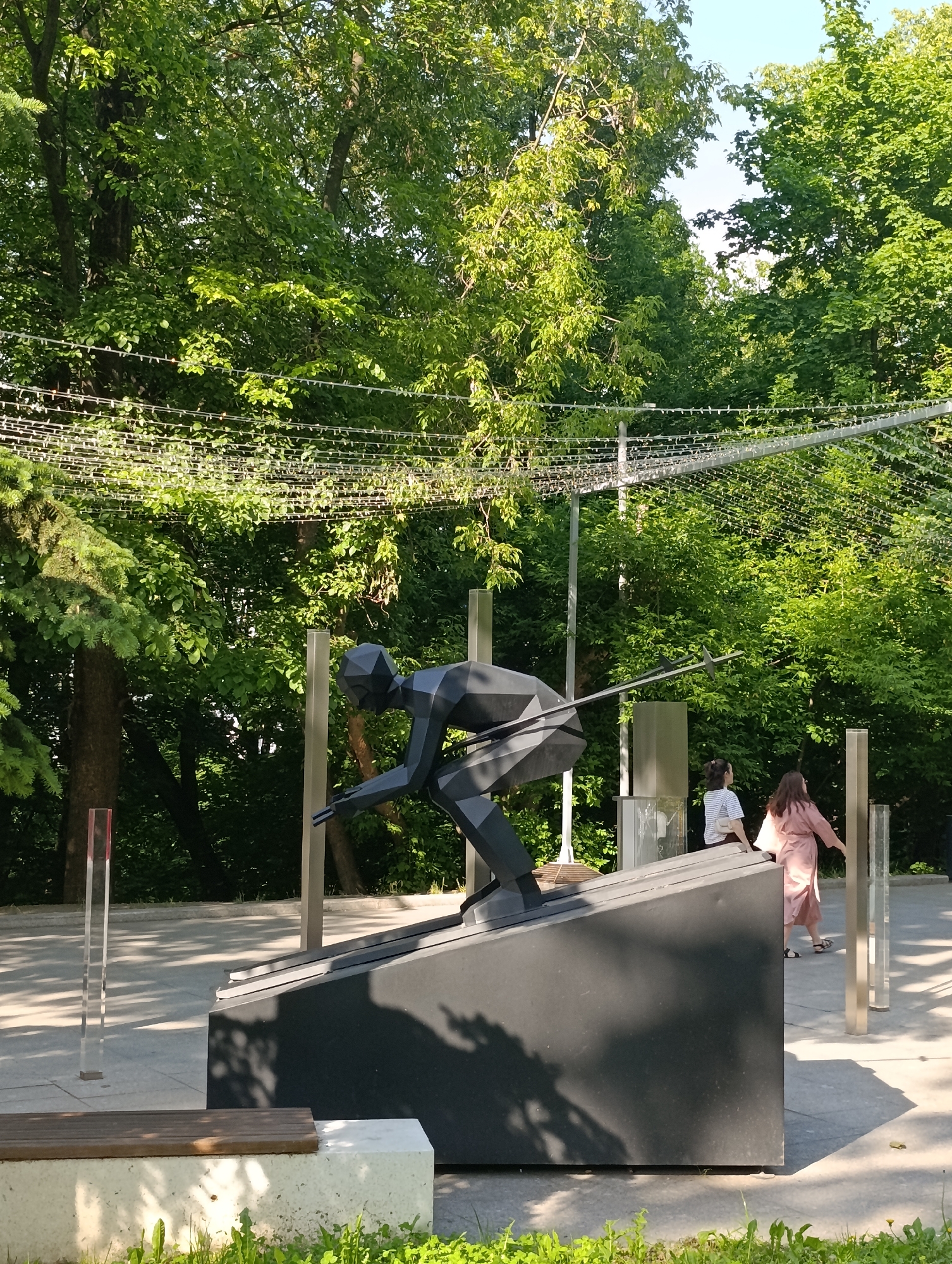 The image displays an outdoor public space, likely a park or plaza, under bright daylight. A large, dark grey, geometric, polygonal sculpture of a biathlete on skis dominates the center-left. The figure is depicted in a crouched, downhill stance, holding a rifle across its body, and is mounted on a matching dark grey, trapezoidal base angled upwards.
In the background, dense green foliage from mature deciduous trees fills the upper two-thirds of the frame, indicating a verdant environment. A network of white wires or decorative lights is strung horizontally across the mid-ground, suspended between poles, creating a canopy-like effect. Sunlight strongly illuminates the scene, casting distinct shadows on the ground and the sculpture.
To the right of the sculpture, two women are present. One woman, wearing a light-colored striped top and dark pants, carrying a light bag, is partially visible behind the sculpture's base. The other woman, dressed in a long, light pink garment and sandals, walks away from the sculpture towards the right side of the frame.
The foreground features a light grey paved surface. On the far left, a wooden bench with a concrete base is visible. Patches of green grass with small yellow flowers are present in the immediate foreground. Several tall, clear or reflective vertical posts are positioned around the sculpture and in the mid-ground, with one on the far left having a red cap. The location is in the city of Unknown, Unknown.