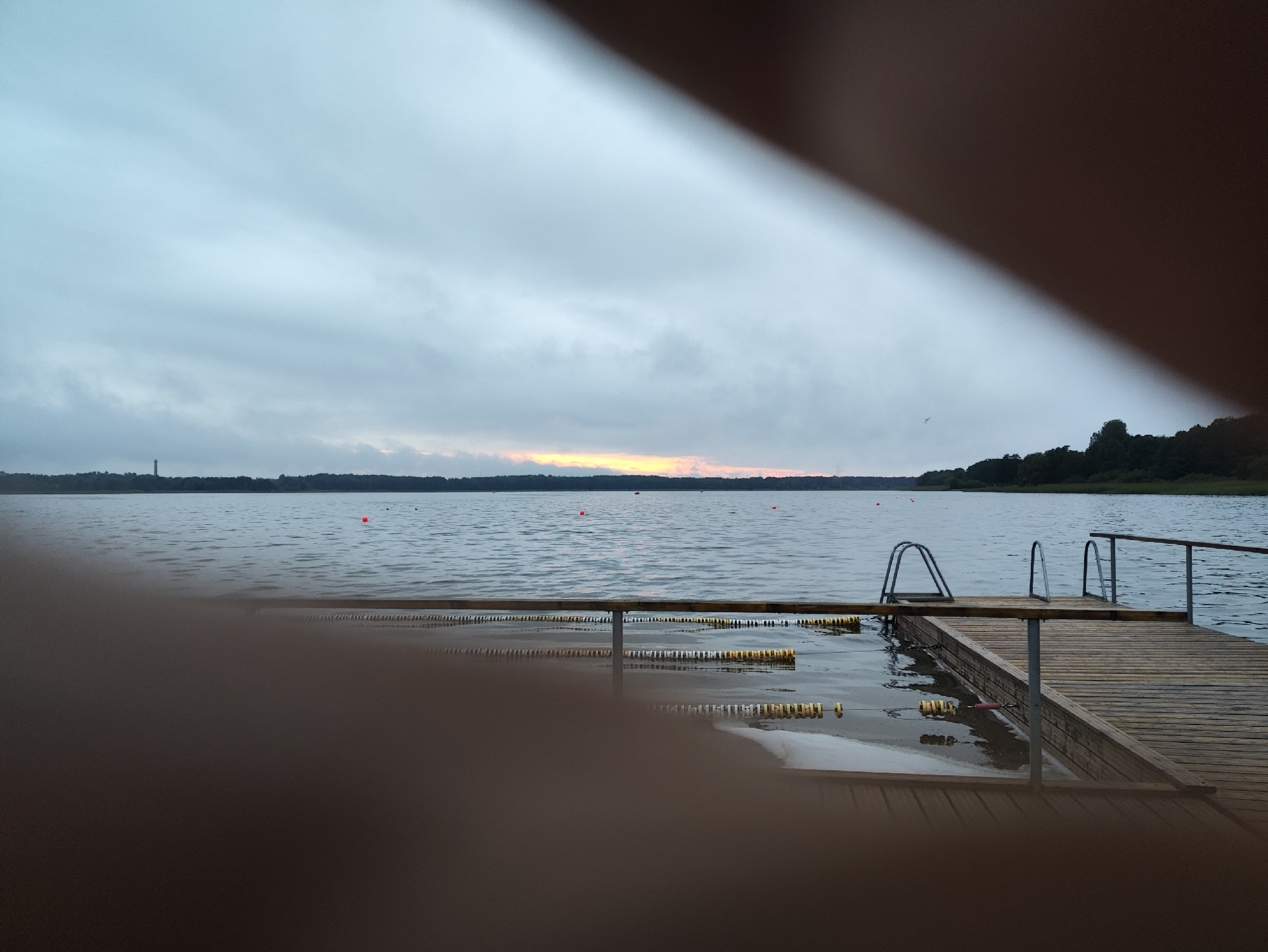 The image captures a serene lake scene at dusk. A wooden pier extends into the calm water, complete with metal railings and ladders for entry. Yellow buoys line a swimming area, and red buoys are scattered further out in the lake. The distant shoreline is densely forested, and a tall, slender structure, possibly a lighthouse or tower, is visible against the hazy sky. The sky itself is overcast with a subtle, warm glow along the horizon, suggesting the last rays of sunlight. The foreground is partially obscured by a blur of brown, likely fingers or hands, indicating the perspective from which the photo was taken. The overall atmosphere is peaceful and quiet. This scene is located at the Pirita beach area in Tallinn, Estonia.