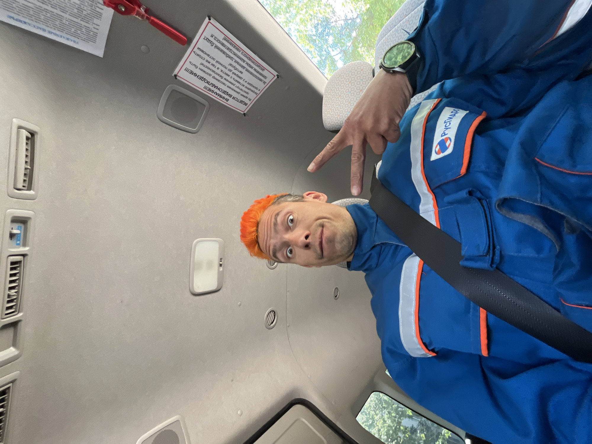 The image is taken from the interior of a vehicle, possibly a bus or van, with the camera facing upwards and slightly to the side. A person with bright orange hair is visible, wearing a blue uniform with white and orange stripes, and a black seatbelt. The person is making a peace sign with their fingers. The uniform has the word "РУСГИДРО" (RUSHYDRO) on it. A watch is visible on the person's wrist. A sticker with Russian text is affixed to the interior roof of the vehicle. The text warns passengers about video surveillance and the requirement to wear seatbelts. Additional text warns against standing or moving in the bus until it comes to a complete stop, noting that failure to comply will result in administrative or disciplinary actions. The roof of the vehicle is light gray and has air conditioning vents and a light. The location is implied to be the city of Chaikovsky, Russia, based on the uniform patch.