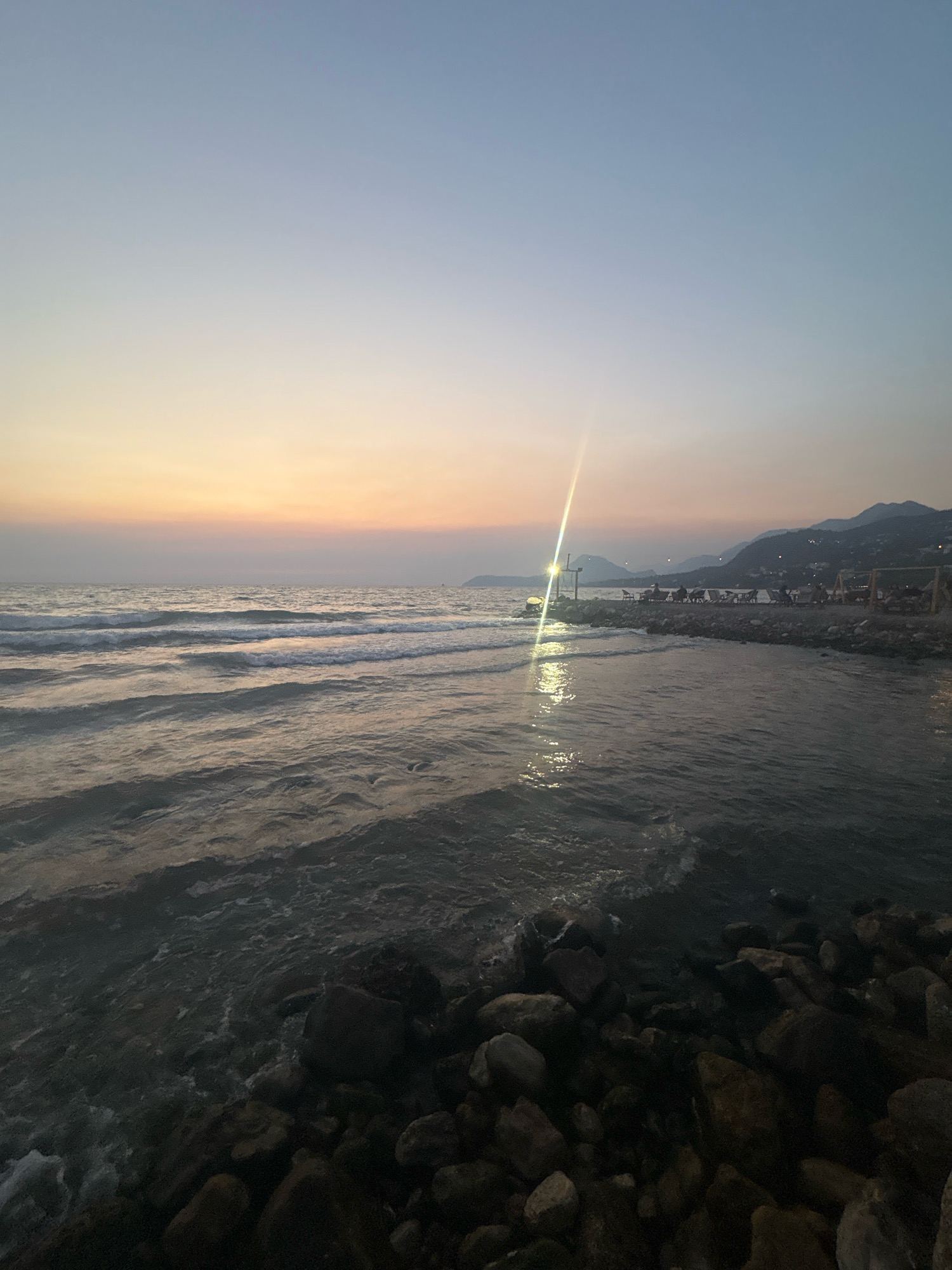The image captures a serene sunset scene at a rocky shoreline in Bar, Montenegro. The sky transitions from a pale blue at the top to soft hues of orange and yellow near the horizon, indicating the sun has recently set. Gentle waves lap against the shore, which is covered with dark, varied-sized rocks.
In the mid-ground, a man-made structure resembling a pier or breakwater extends into the sea, lined with lounge chairs and a few people are seated. A bright light source, possibly a lamp post, is prominently visible on this structure, reflecting intensely on the water's surface. Beyond the shoreline, a silhouette of hills or mountains forms the background, with scattered lights suggesting a town or settlement nestled within them. The overall atmosphere is tranquil and peaceful, typical of a coastal evening.