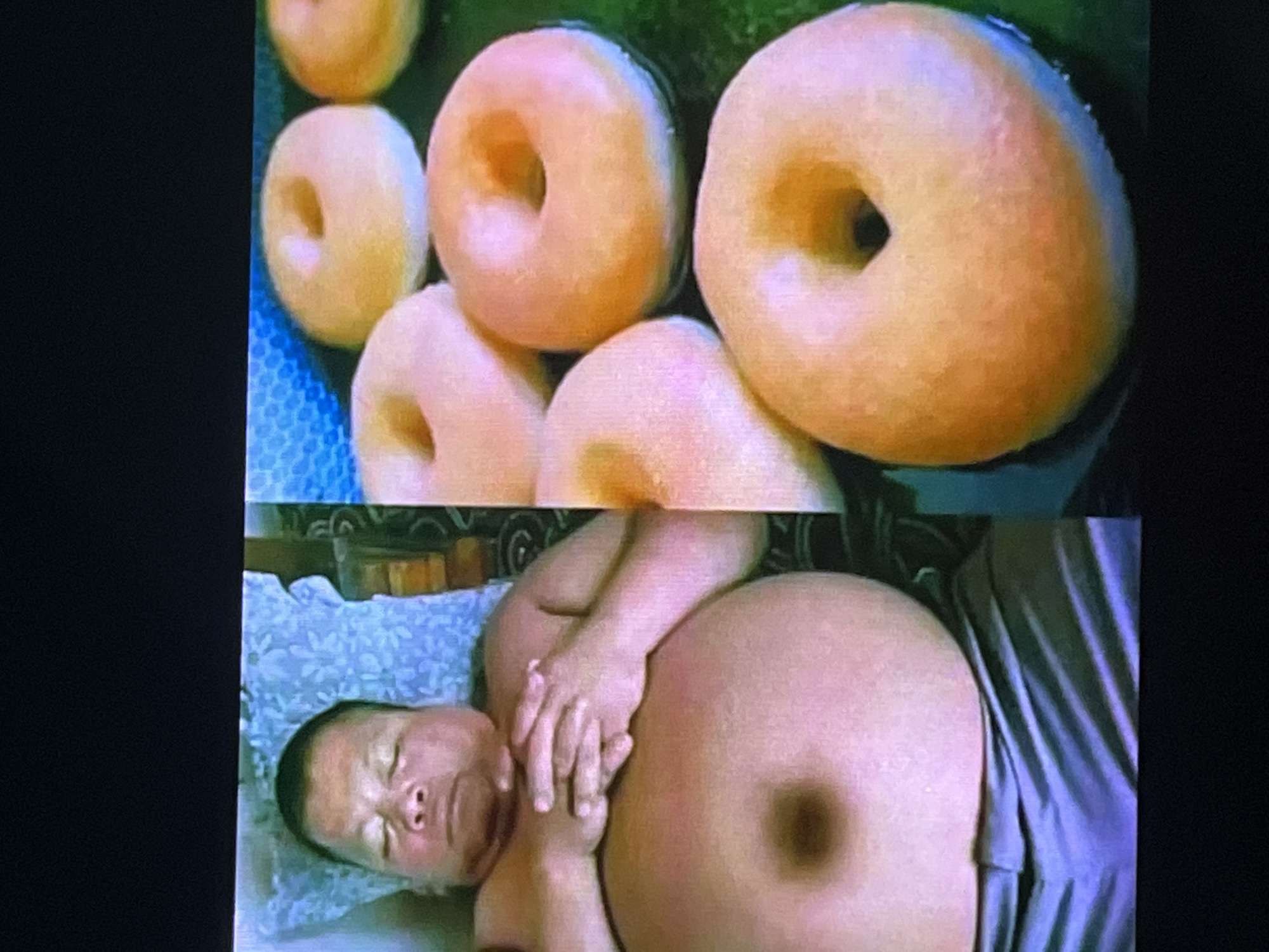 The media file contains two distinct images, one above the other, separated by a horizontal line. The top image displays a close-up view of several plain, ring-shaped donuts. They are a light golden-brown color, with a slightly pinkish hue, and are arranged in a slightly overlapping manner. The donuts appear to be on a dark, possibly green, surface, with a glimpse of a blue, textured object on the left side.
The bottom image features a baby lying on its back, with its eyes closed and a serene expression. The baby is positioned on a light blue, patterned cloth. An adult's hands are visible, gently holding the baby's head and resting on its chest. To the right of the baby is a large, round, pale pink object, which appears to be the abdomen of an adult wearing a light blue garment. The lighting in this scene suggests an indoor setting, possibly during the daytime.
There is no visible text in either image. The overall impression is a juxtaposition of food items with an intimate moment between a baby and an adult.