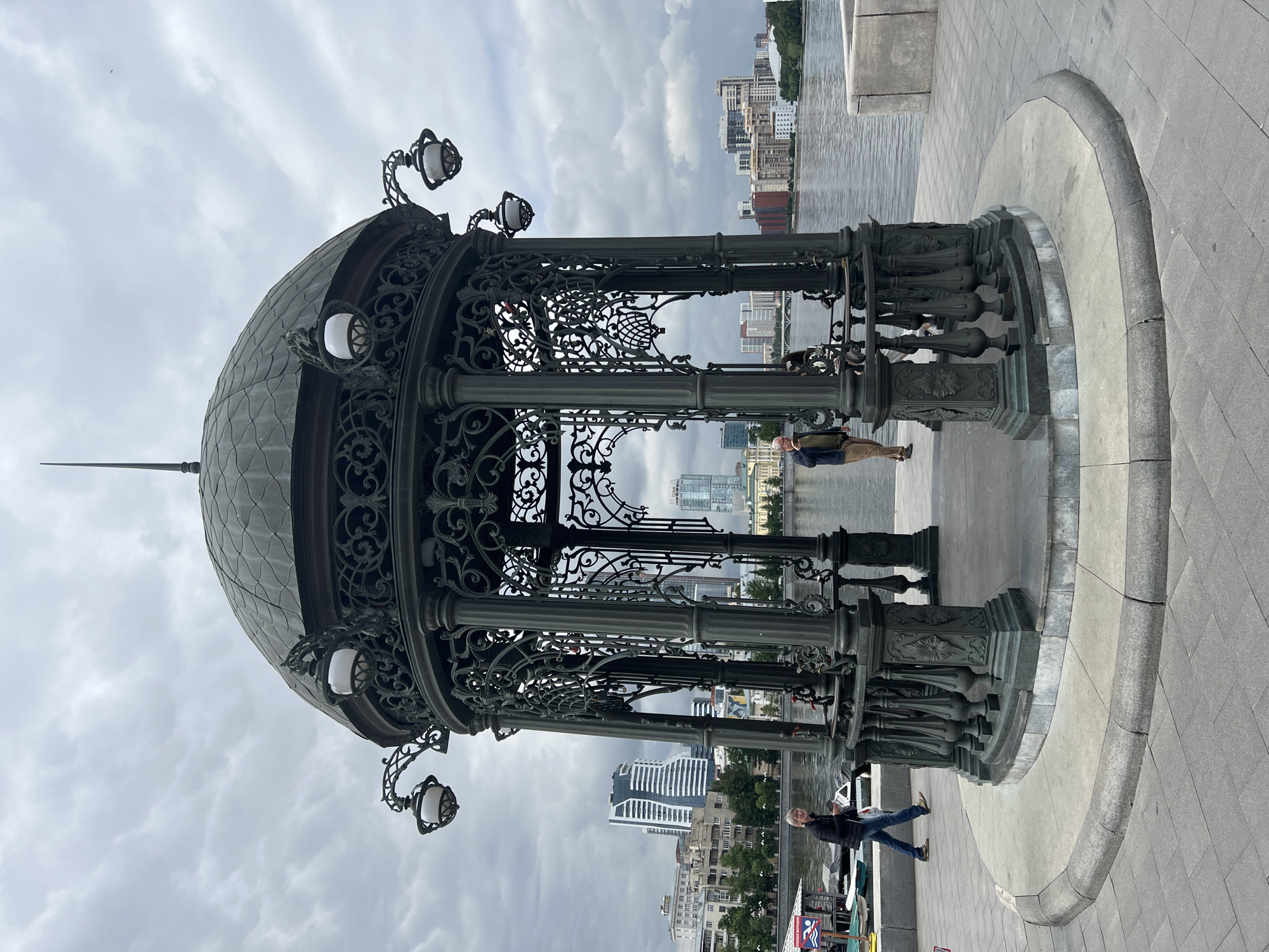 A dark green, ornate gazebo stands on a paved walkway next to a body of water. The gazebo has a domed roof with a finial and four lampposts extending from its sides. The structure is made of wrought iron with intricate scrollwork. Two people are visible within the gazebo; one man is standing and looking out at the water, while another woman is walking past on the walkway to the left. In the background, across the water, is a cityscape with modern buildings and trees. The sky is overcast with gray clouds. The scene is located in Yekaterinburg, Russia, on the embankment of the Iset River.