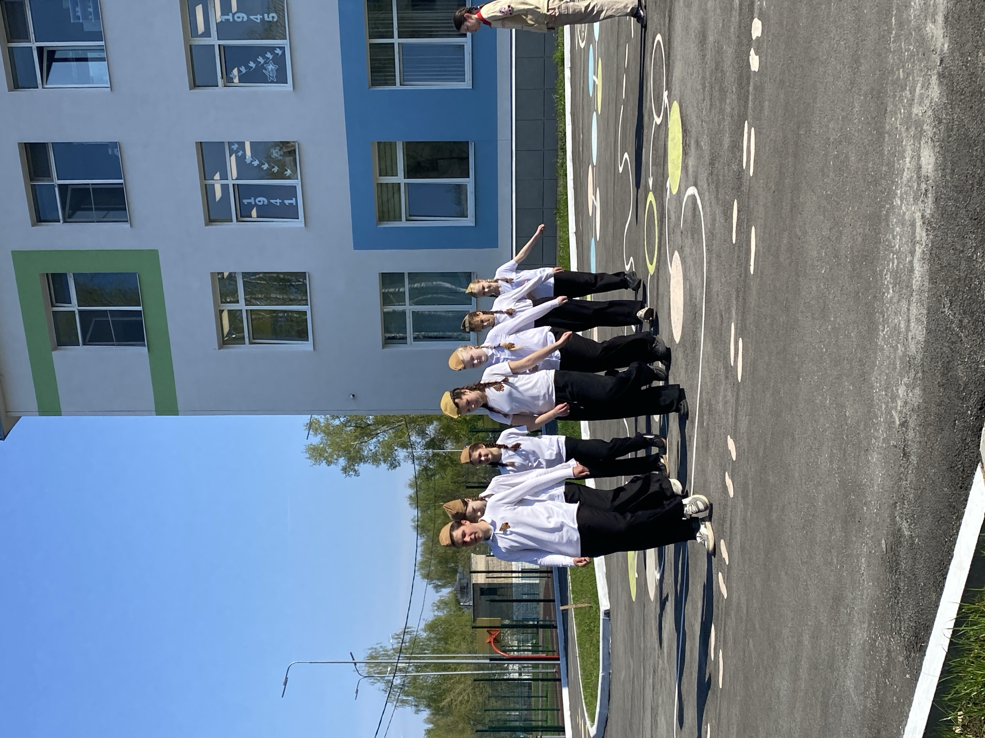 The image shows a group of children standing at attention on what appears to be a playground or asphalt surface marked with hopscotch patterns. They are dressed in white shirts and black pants or skirts, with hats of a light brown color. A single person, seemingly an adult, in a beige uniform with a red neckerchief stands at the head of the line. The background includes a building with windows decorated with "1945" markings and dove designs, suggestive of a commemorative or celebratory theme, likely related to Victory Day marking the end of World War II in Europe. A lamp post and trees are visible beyond the group. Based on the signage, the location is identified as the city of Chaikovsky, Russia.