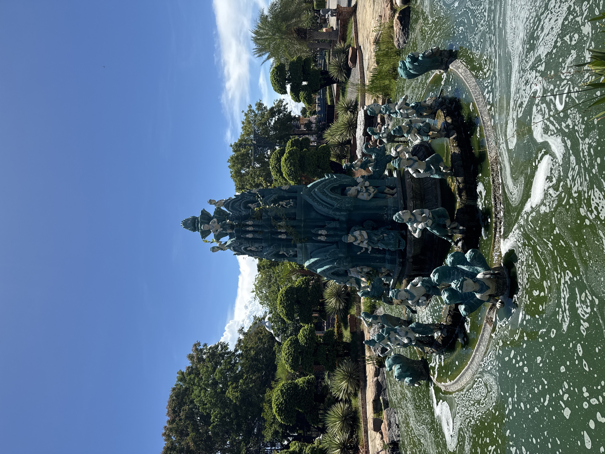 A tall, ornate, greenish-blue sculpture/fountain structure resides in a body of greenish water, with a foam-like substance floating on its surface. The sculpture appears to be multi-tiered and adorned with human figures. Around the water feature, there are landscaped areas with trees and plants. The sky is blue with some clouds. It is located in the city of Bang Pu Subdistrict Municipality, Thailand.