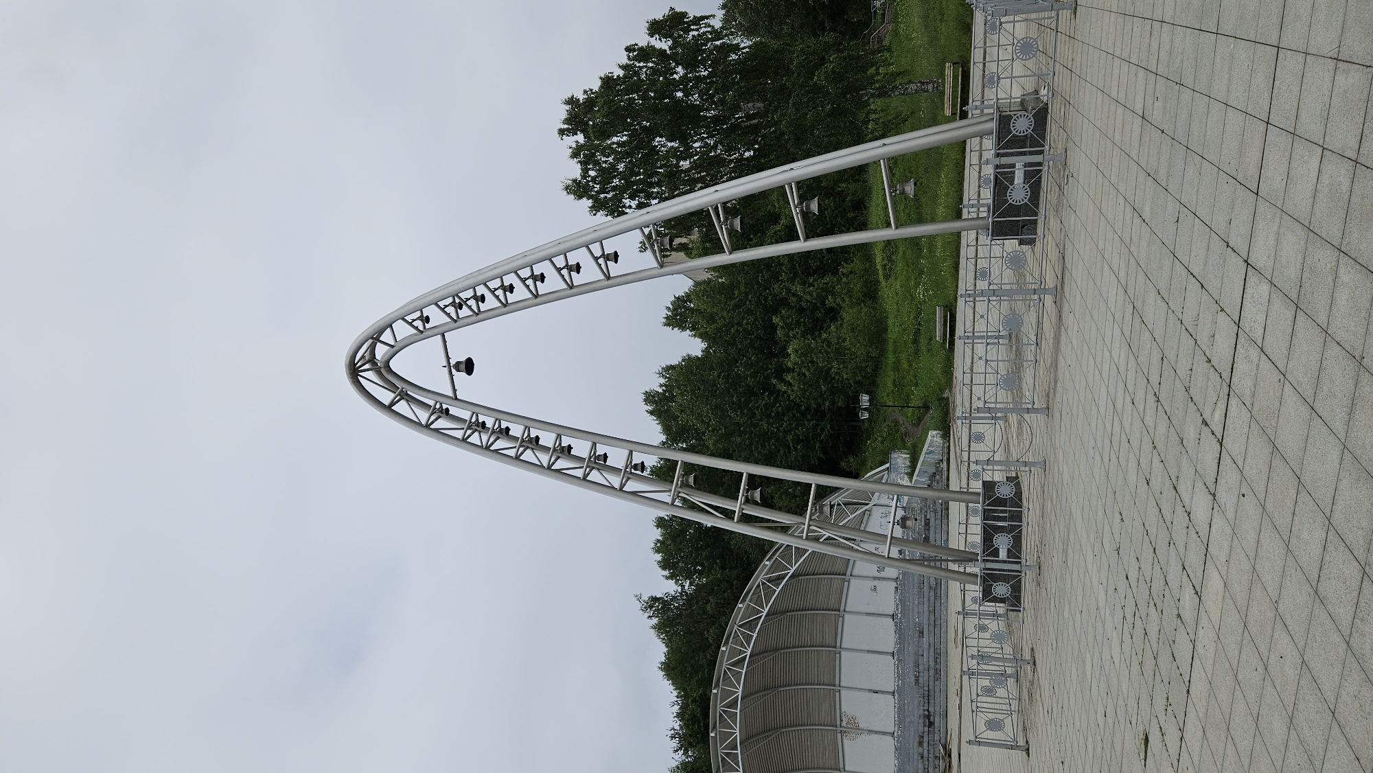 An outdoor shot captures a large, modern, arch-shaped metal structure adorned with numerous small bells suspended from its frame. The structure stands in a paved, open area, with a decorative metal fence encircling its base. Behind the structure, a lush green hillside is covered with trees. To the left, a portion of a white, amphitheater-like seating area is visible. The sky is overcast and gray.