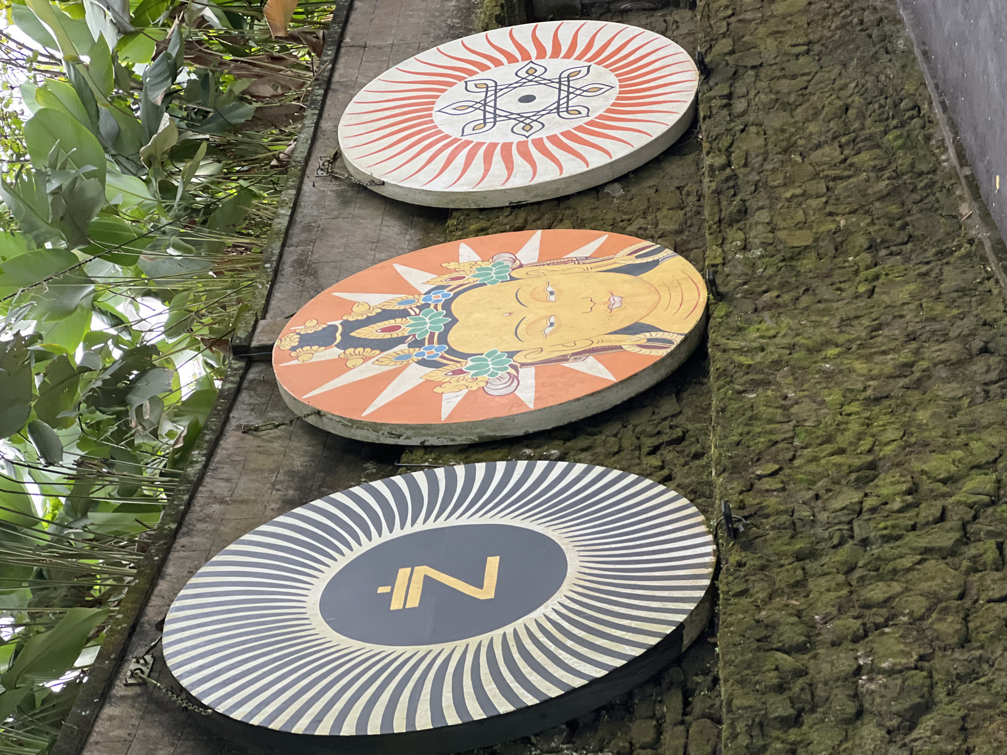 The image shows three circular signs mounted on a textured, moss-covered wall. Green foliage is visible to the left of the signs. The top sign features a white background with a red radiating pattern and a black geometric design in the center. The middle sign has an orange background with a yellow face, a floral headdress, and a radiating pattern. The bottom sign is dark gray with a gold-colored "IN" symbol in the center and a white radiating pattern around it. The setting is a wall possibly in the Ubud District, Indonesia.