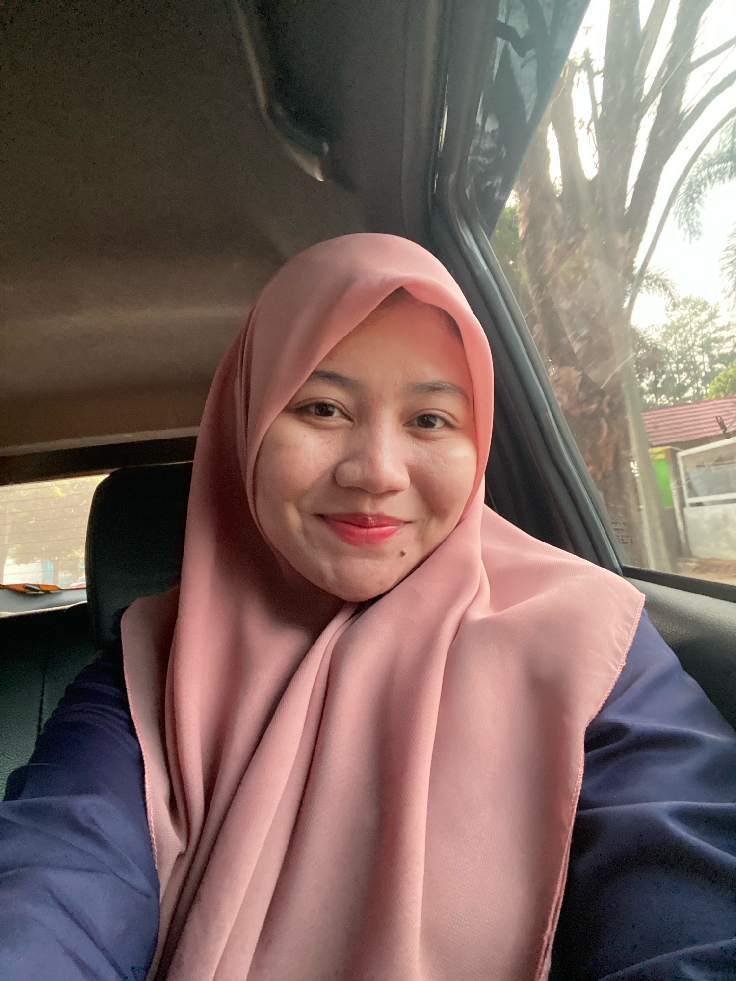 A young woman wearing a pink hijab and a dark blue top is seated in the back of a vehicle, possibly a car or van. She is looking directly at the camera and smiling, with red lipstick on her lips. Her right cheek shows a dimple, and a small mole is visible below her lower lip on the left side of her face. The background outside the vehicle's window includes trees and a building with a red roof, suggesting an outdoor, possibly suburban, environment. The lighting suggests it is daytime. The interior of the vehicle shows a dark seat and the car's door frame. The scene appears to be in Banjarbaru, Indonesia, given the context of the location provided.