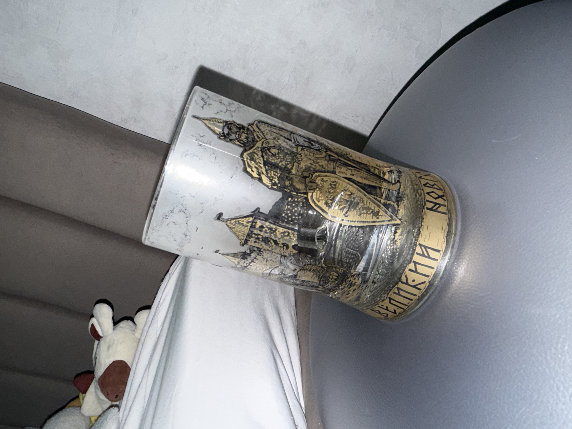 The image shows a glass tumbler with a gold-colored design depicting a historical figure in armor with a shield and a depiction of a castle or fortified city. The inscription around the base of the glass appears to be in Russian Cyrillic script. The glass is positioned on a soft surface, possibly a chair or bed. To the left, there's a stuffed animal. The environment suggests an indoor setting. The tumbler depicts historical scenes related to the city of Veliky Novgorod, Russia.