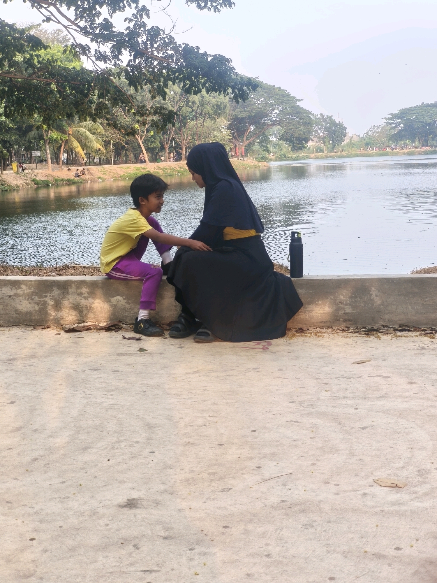 In a park beside a body of water in Bekasi, Indonesia, a young boy wearing a yellow t-shirt and purple pants sits on a concrete ledge, facing a woman in a black hijab and long dress. The woman is reaching out to the boy, and they appear to be interacting. A black water bottle rests on the ledge behind the woman. The background shows trees and more of the park, with a cloudy sky overhead. The ground in the foreground is a rough concrete surface with some leaves and debris.