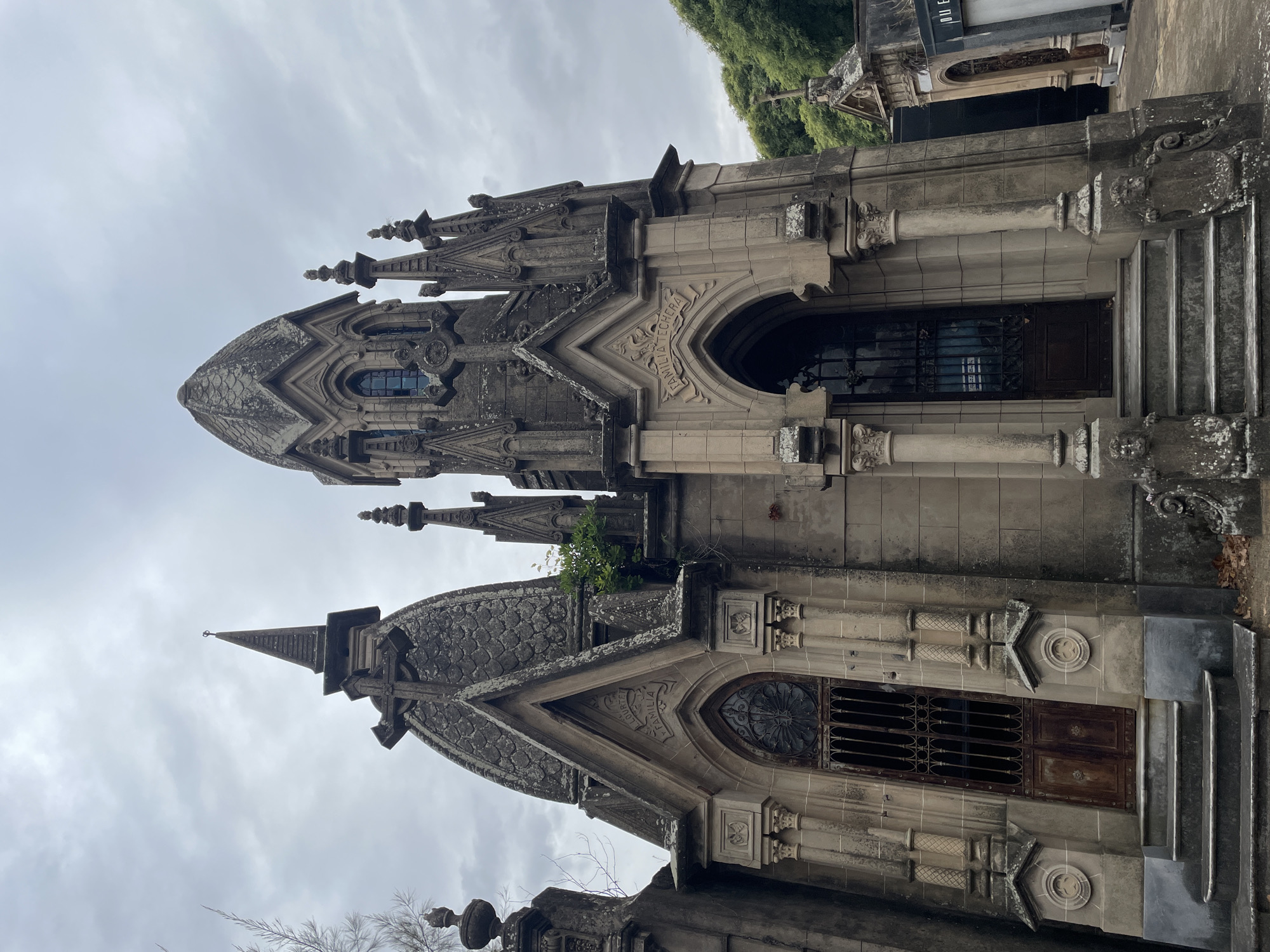 This image captures a section of the Recoleta Cemetery in Buenos Aires, Argentina, under an overcast sky. The scene features two intricately designed, weathered stone mausoleums, constructed in a Gothic Revival architectural style, facing slightly towards the viewer.
The mausoleum on the left is characterized by a pointed, scaled stone roof culminating in a finial topped with a cross, and a rounded, scaled dome positioned behind it. Its facade includes a prominent pointed arch entrance, fronted by a heavily rusted, dark metal gate with decorative elements, and a solid wooden door set back. Above this arch, the name "FAMILIA LOURTEL" is carved into the stone.
Immediately adjacent on the right, the second mausoleum displays a rounded, ribbed stone dome with multiple narrow, arched windows, some appearing dark blue or reflective. This structure is adorned with multiple spires and finials. Its main entrance is also a pointed arch, featuring a dark metal gate and a wooden door. Above this arch, the inscription "FAMILIA TECHERA" is visible. Both structures exhibit detailed stone carvings, decorative columns, and pilasters. Multiple stone steps lead up to each entrance. The stonework throughout is discolored by age and environmental exposure, with visible patches of moss and lichen.
Sparse, bare tree branches are visible in the upper left corner, while dense green foliage, likely from trees, frames the background on the far right. Additional, smaller mausoleums are partially visible in the background, further establishing the cemetery setting. Dried leaves are scattered on the ground near the base of the structures. No individuals, actions, or interactions are depicted in the scene.