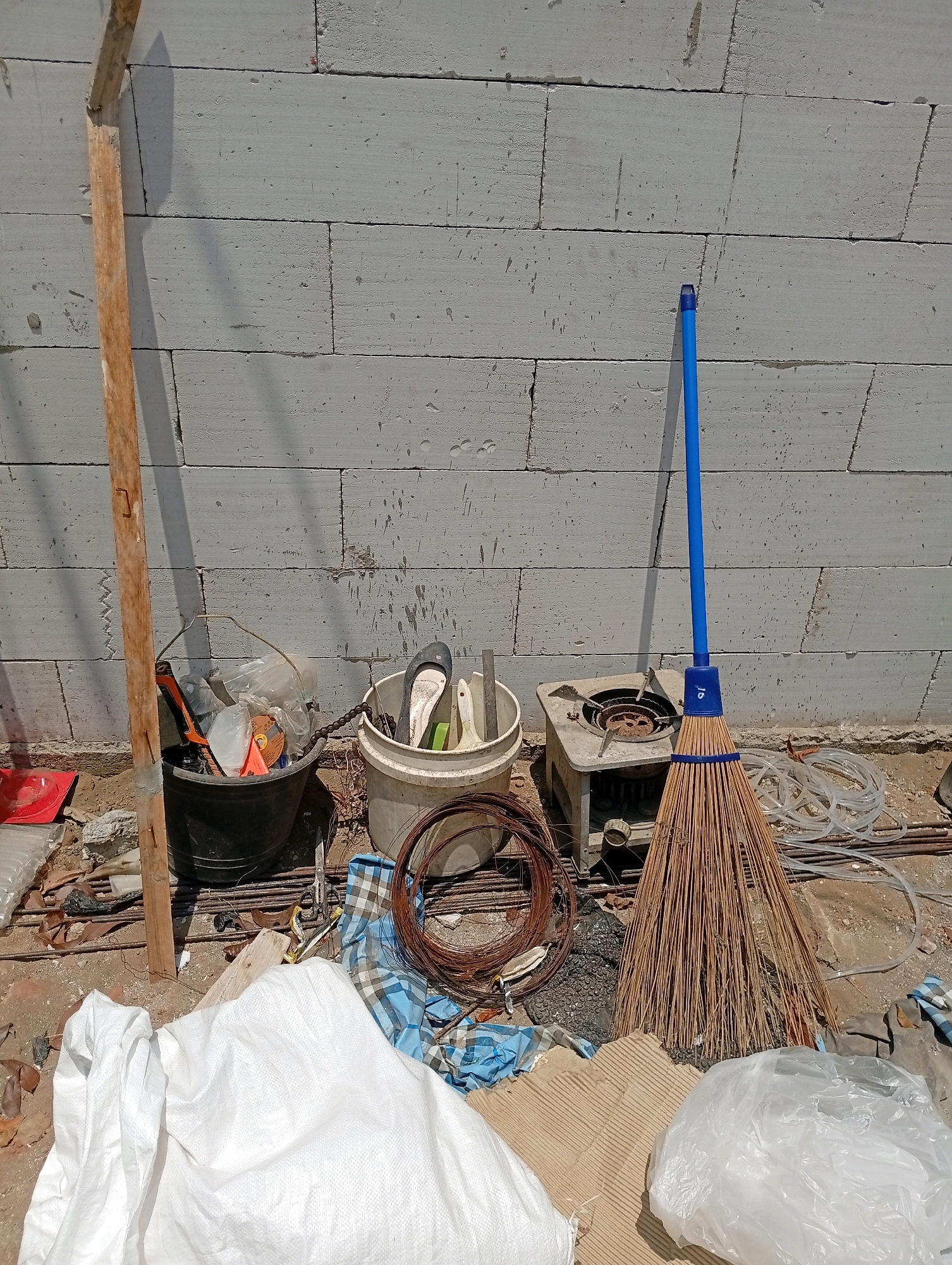 The image depicts an outdoor construction site in Blimbing, Indonesia, under bright sunlight. In the foreground, a large white woven bag and a smaller white plastic bag are visible, lying on a surface of dirt and debris. To the left, a weathered wooden plank is propped against a wall made of pale gray, rectangular blocks. A black plastic bucket filled with various construction tools, including what appears to be an orange utility knife and plastic bags, sits near the plank. Adjacent to this, a white bucket contains a pair of black and white shoes, a green object, and a white brush. Further to the right, a small, metallic object resembling a portable stove or burner is present. Dominating the right side of the frame is a broom with a blue handle and natural bristles, leaning against the block wall. Various construction materials, including coiled wire and clear plastic tubing, are scattered on the ground behind the bucket and broom. The wall behind the objects is made of large, light gray cinder blocks, with visible mortar lines and some dark smudges. The ground is uneven and covered with dirt, small stones, and debris.