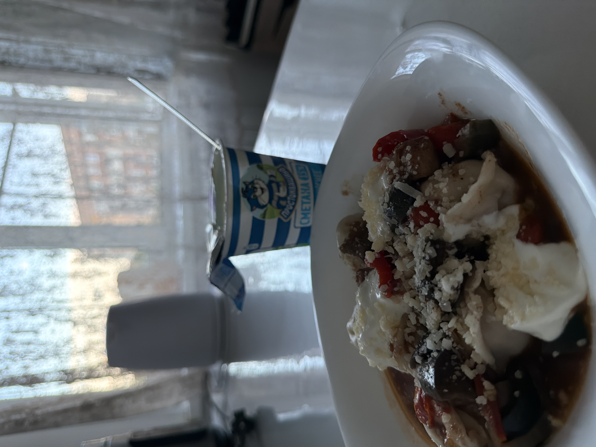 The image shows a white bowl containing food, likely a salad, with visible ingredients like eggplant, red peppers, and a white sauce or dressing, possibly topped with grated cheese. A partially opened container of "Smetana" (sour cream in Russian) is positioned beside the bowl with a spoon sticking out of it. The container's packaging features blue and white stripes and a cartoon cat. The background includes a blurred window and a white countertop. The image appears to have been taken indoors, potentially in Leningradskaya, Russia, based on the packaging of the sour cream.