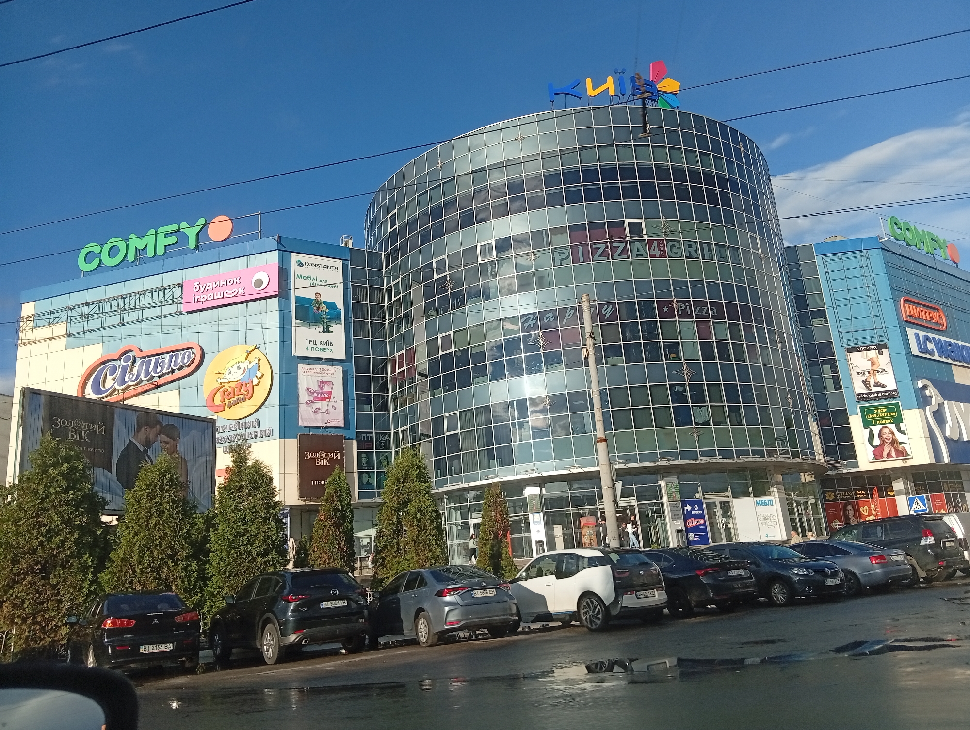 The image depicts the exterior of a multi-story shopping center in Poltava, Ukraine. The facade features large signage for "COMFY," "Silpo," "Pizza4Grill," and "Kiyv." There are advertisements for furniture, toys, and jewelry. A row of parked cars is visible in front of the building, separated by several evergreen trees. The sky is blue, and the scene appears to be in daylight. The street is wet, suggesting it has recently rained. There is a pedestrian area in front of the building where individuals are walking. Electrical wires are visible above the building.