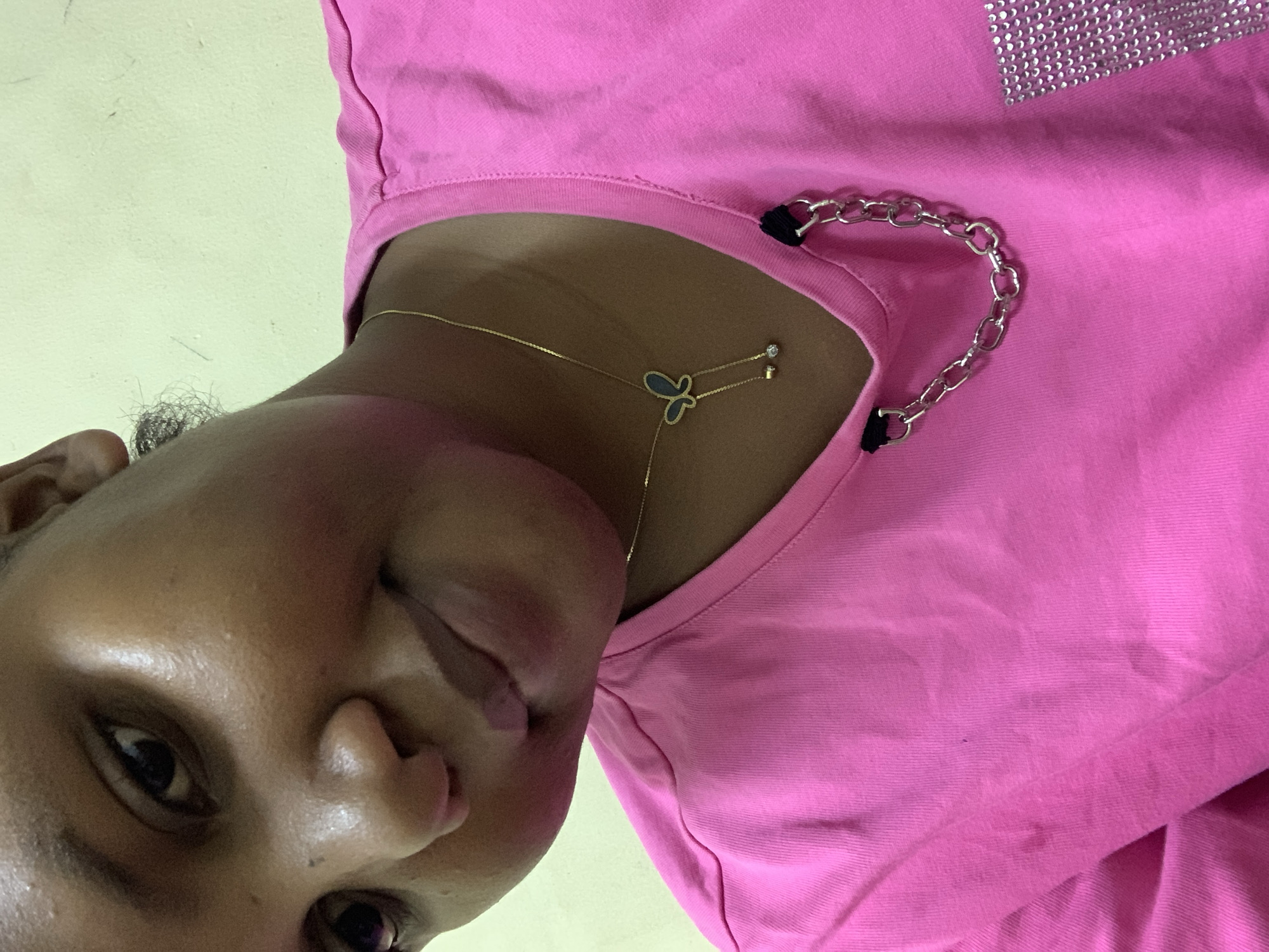 A close-up, angled selfie of a dark-skinned person wearing a pink top with a silver studded pattern on the upper right and a silver chain detail around the neckline. The person is also wearing a delicate gold necklace with a butterfly-shaped pendant and a small gem. The background appears to be a pale yellow wall. No information from the photo indicates the city is Dar es-Salaam, Tanzania.