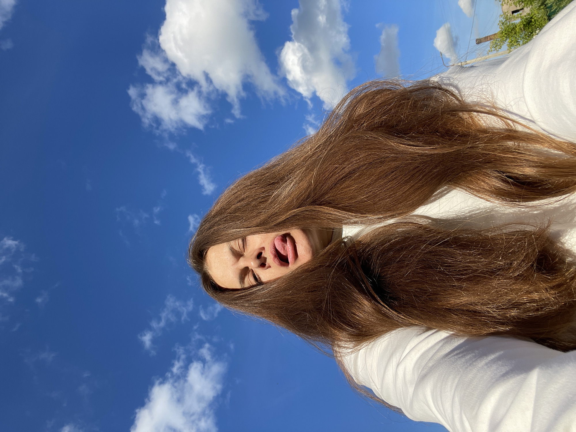 This is a low-angle, close-up, full-body shot of a person outdoors on a bright, sunny day. The sky is a vibrant, clear blue with scattered white, puffy clouds, suggesting fair weather. The person, whose face is visible with their eyes closed and tongue sticking out in a playful expression, has long, brown hair that cascades around them, framing their face and obscuring part of their white shirt. The camera is angled upwards, capturing the vast expanse of the sky and the person from a unique perspective. In the background, a distant tree and a utility pole are visible, hinting at a semi-rural or suburban setting. The sunlight is strong, casting shadows and highlighting the texture of the person's hair and clothing. The image is oriented vertically, with the top of the sky appearing at the bottom of the frame. There is no visible text. The scene conveys a sense of carefree joy and freedom.
