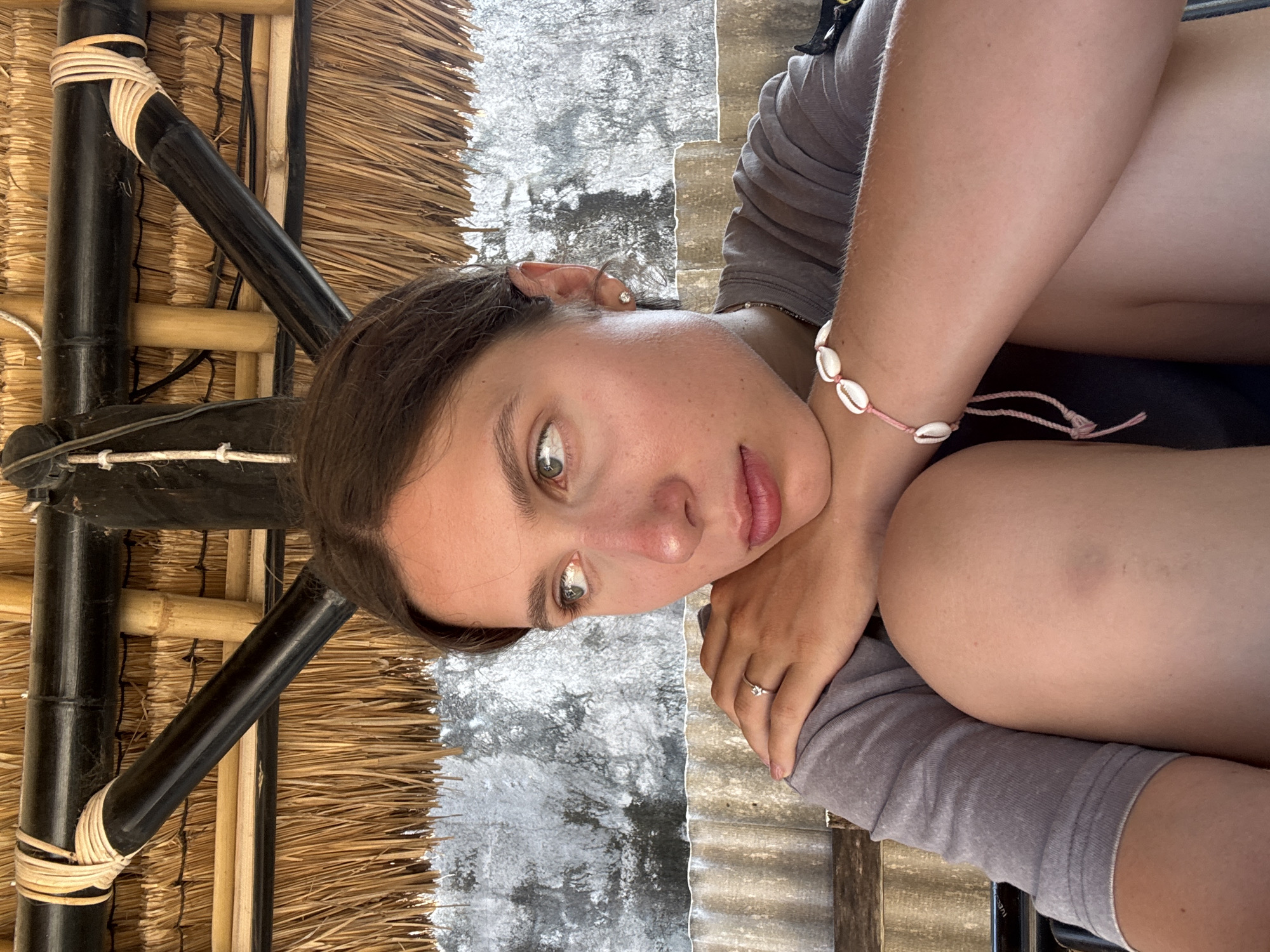 A young woman with light brown hair is seated in an outdoor covered area, looking to her left. She is wearing a grey t-shirt and a bracelet made of small white shells on her right wrist. A ring with a small stone is visible on her left hand, which is resting on her shoulder. The background shows a thatched roof supported by bamboo poles, a textured concrete wall, and corrugated metal sheeting. The setting appears to be Kertasari, Indonesia, possibly a beachside hut or cafe, given the tropical materials and the woman's casual attire.