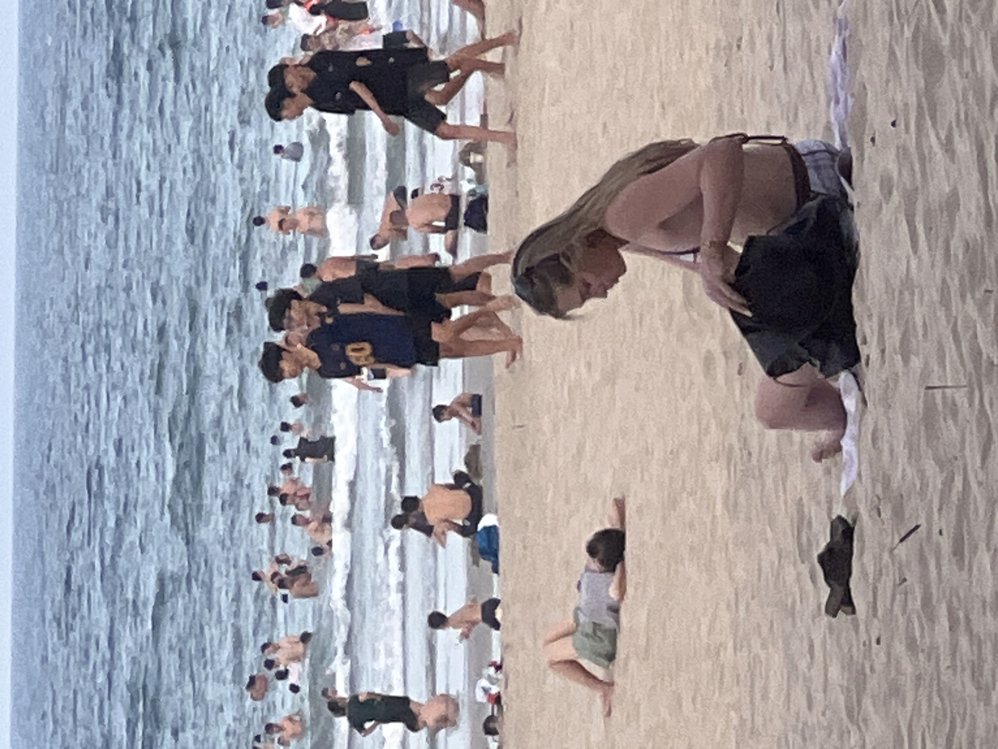 This media file captures a crowded beach scene in Đà Nẵng, Vietnam. The image is oriented vertically, presenting a view of the ocean on the left and the sandy beach on the right.
The ocean is a prominent feature, with waves gently rolling towards the shore. Numerous people are visible in the water, some appearing to be wading or playing near the edge, while others are further out. The water has a bluish-gray hue, suggesting it might be overcast or that the water is not particularly clear.
The beach is populated with many individuals enjoying the seaside. On the sand, a woman with long, light-brown hair is leaning forward, seemingly attending to something near her feet. She is wearing a bikini top and patterned shorts. Further down the beach, other people are scattered, some sitting or lying down, while others are standing. The sand is a light beige color, marked with footprints and natural textures.
The crowd suggests a popular time for beachgoers, likely during daylight hours. The weather appears to be somewhat overcast, as there are no strong shadows visible, and the overall lighting is soft. The presence of many people in swimwear indicates a recreational setting, possibly a tourist area given the location context of Đà Nẵng. There is no visible text in the image.