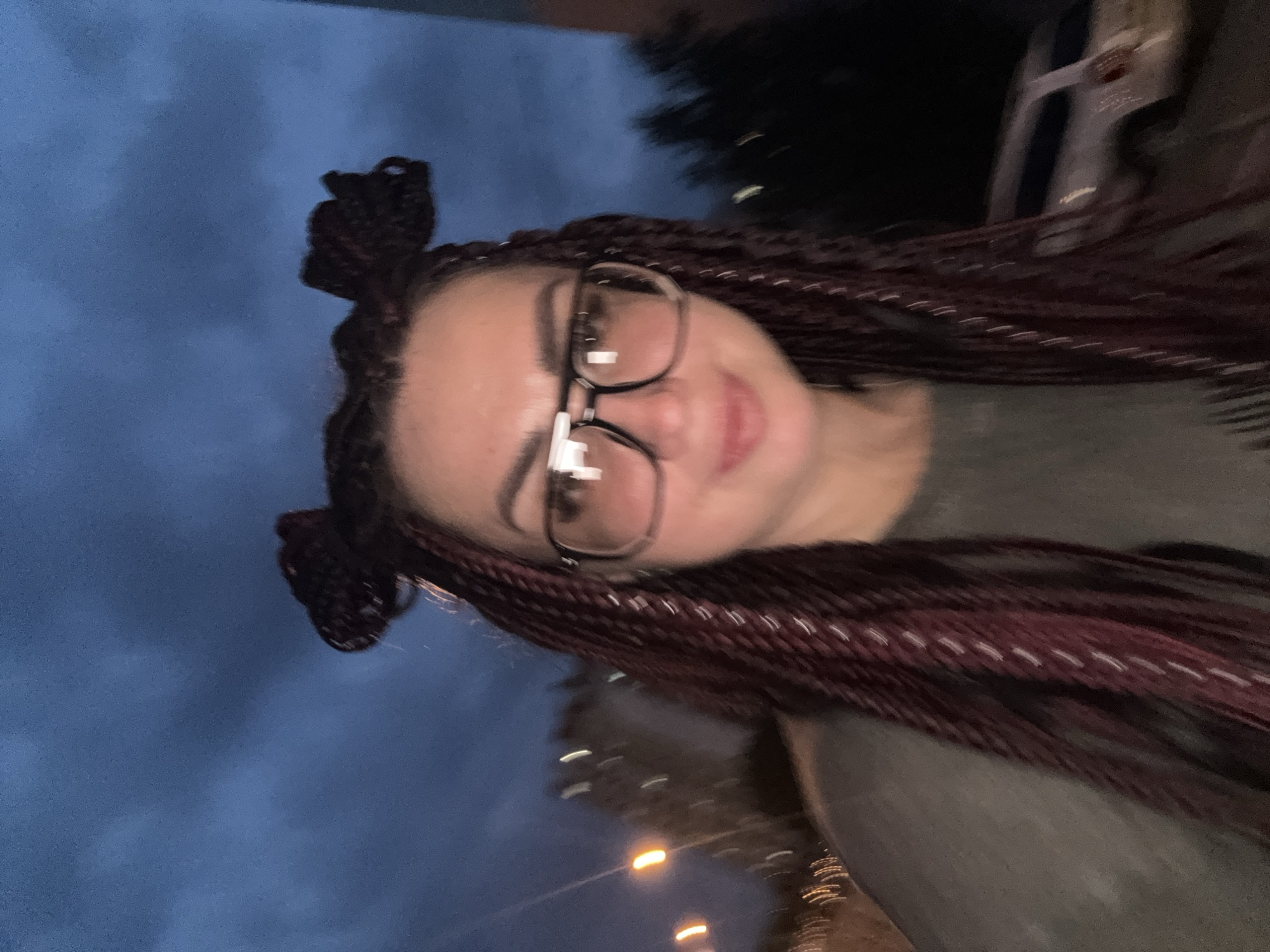 A slightly blurred, low-light, medium shot captures a woman with dark braided hair styled in two buns on either side of her head. She is wearing large, rectangular, dark-rimmed glasses and a dark olive-colored shirt. The background is a deep blue sky with indistinct cloud formations. To the left of the woman, out-of-focus lights suggest streetlights or building lights. In the far right background, the blurry outline of a white vehicle is visible. The image appears to be taken outdoors in an urban environment, possibly Tolyatti, Russia, given the provided context.