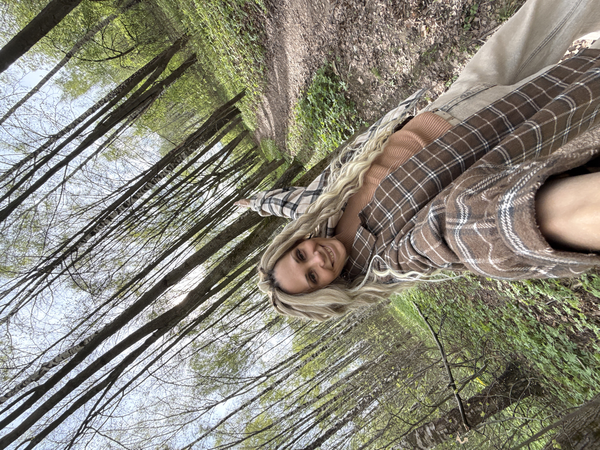 A woman is standing in a forest, possibly located in Rusanovo, Russia. The woman has long, blonde hair and is wearing a brown and white plaid shirt over a tan shirt. She is smiling and extending her right arm forward. The forest consists of tall, thin trees, with a dirt path visible in the background. The sky is visible through the branches. The woman appears to be standing among green undergrowth.
