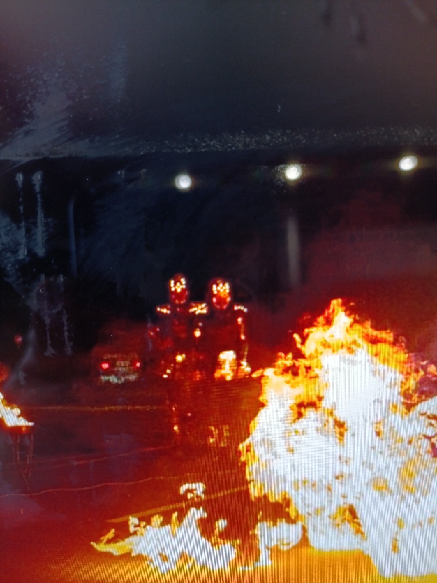 The image depicts a nighttime scene with two figures standing in front of a large bonfire. The figures appear to be wearing some kind of protective gear or costumes with illuminated elements on their heads and chests. They are positioned to the left of a vibrant, brightly burning fire that dominates the right side of the frame. In the background, there are some blurry lights and shapes, possibly indicating a street or an urban environment. A vehicle with red taillights is visible behind the figures, suggesting the presence of traffic or abandoned vehicles. The overall atmosphere is dramatic and somewhat ominous due to the darkness, the intense fire, and the unusual appearance of the figures.