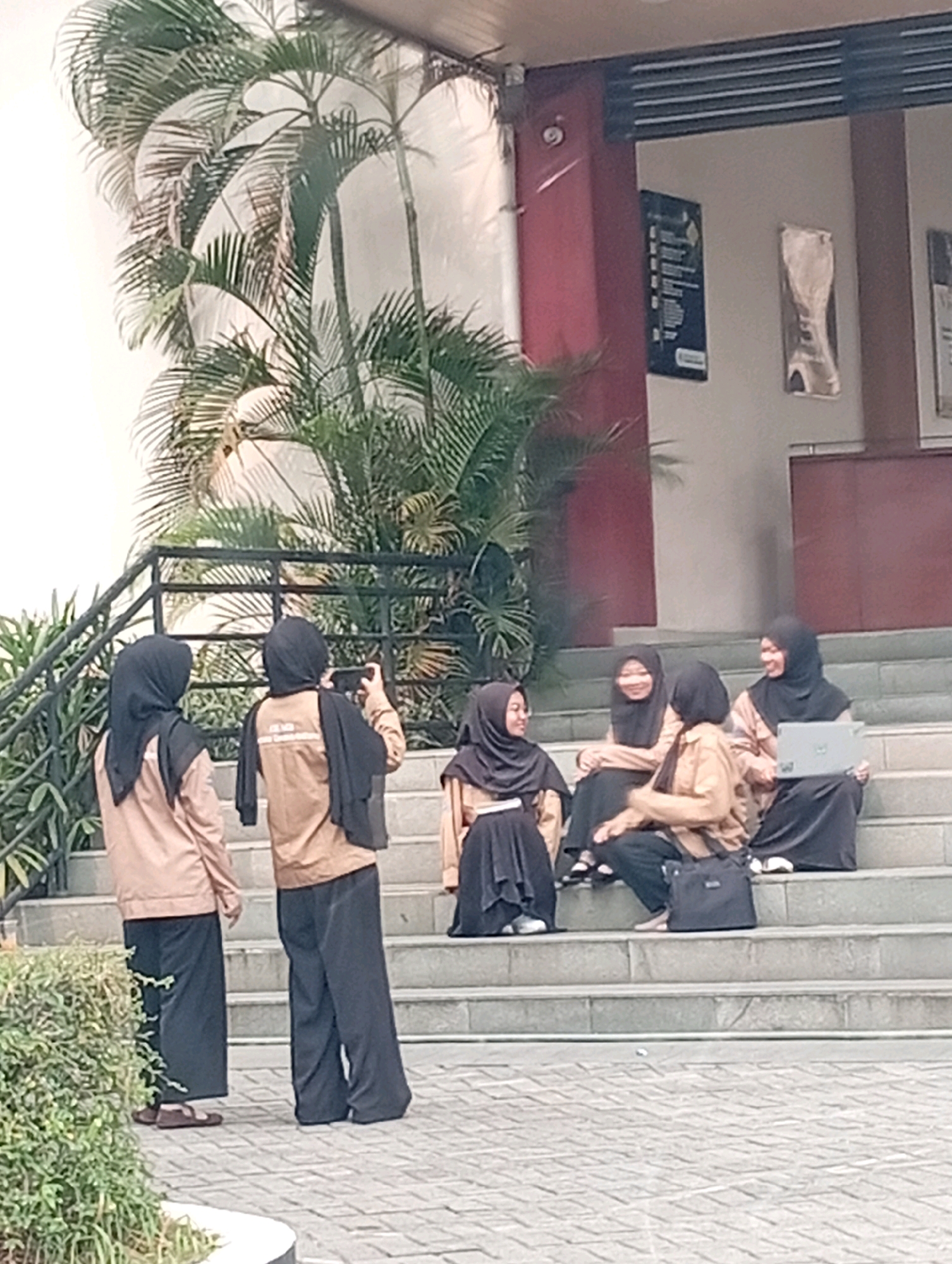 The image depicts a group of five young women, all wearing hijabs and casual attire, some with jackets, gathered on outdoor stairs in front of a building with red accents. The women are positioned in various ways: two are standing, one is taking a picture with a phone, another is holding a camera, and three are seated on the steps. One of the seated women is using a laptop. The environment includes lush green plants, including palm trees, and a paved walkway. The architectural style suggests a modern building, possibly part of an educational institution or public space. The lighting indicates daytime. The scene appears to be set in a relaxed, social atmosphere. The location is identified as Kelapa Dua, Indonesia.