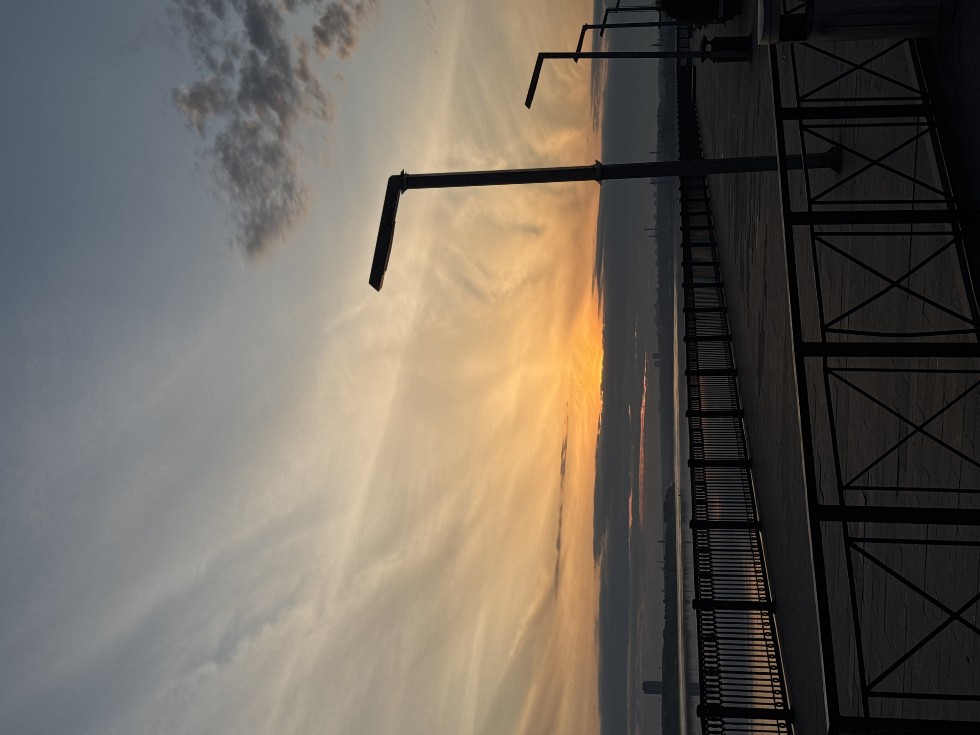 The image depicts a sunset view from the embankment in Perm, Russia. A pedestrian walkway runs along the right side of the frame, defined by a dark railing. Vertical posts with angled street lamps are positioned at intervals along the walkway. A body of water, likely the Kama River, is visible beyond the railing, reflecting the sunset colors. The sky is filled with wispy clouds, colored in shades of yellow, orange, and gray by the setting sun. Distant buildings are barely visible on the opposite bank of the river, obscured by the atmospheric haze. The lighting is generally low, suggesting the photograph was taken during the early evening.