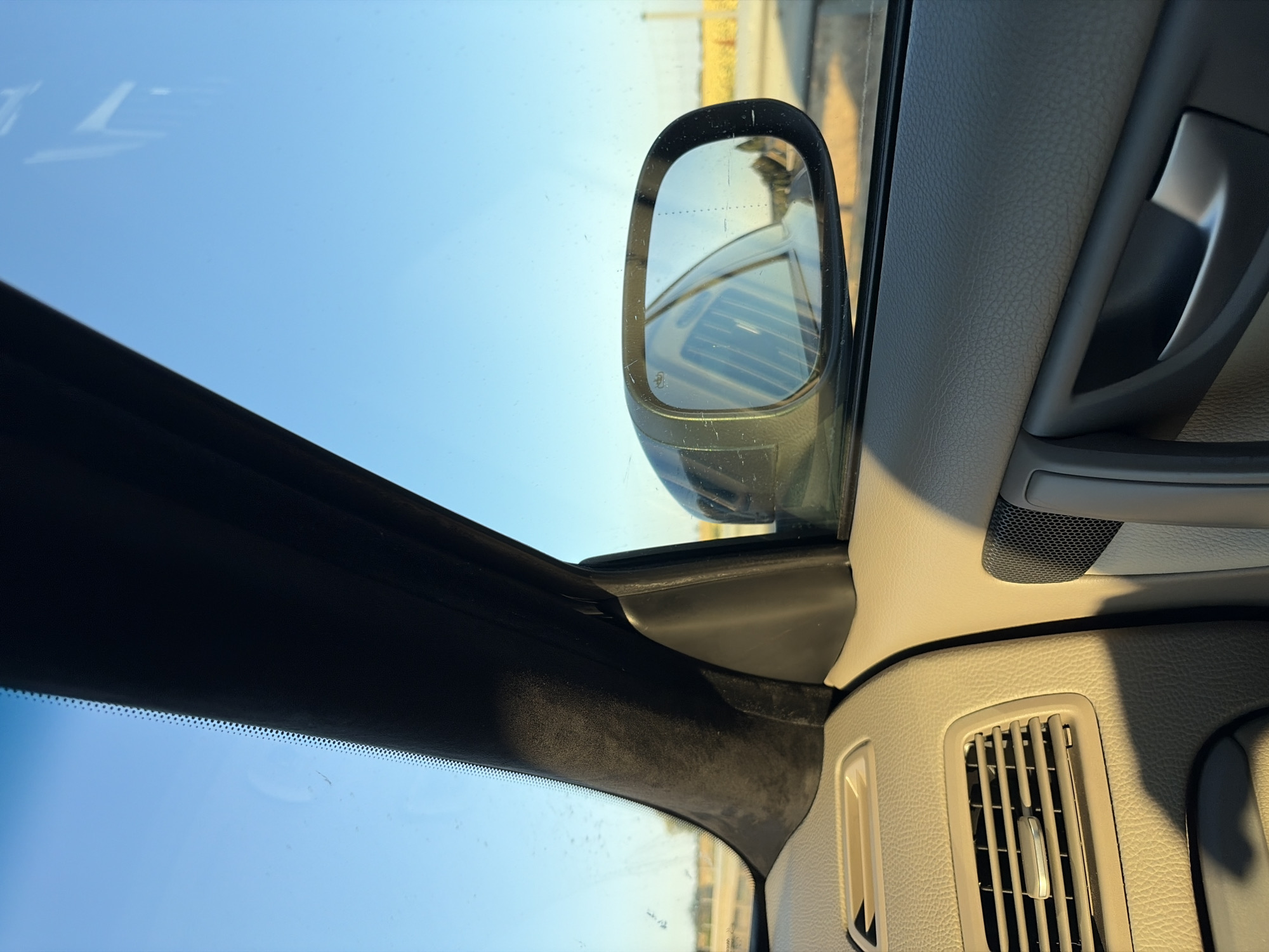 The image depicts the interior of a car, looking out the passenger window and at the side mirror. The dashboard and door panel are visible, along with an air vent. The side mirror reflects the sky and a landscape of fields, suggesting the car is on a road. The sky is clear and blue, and the sun is casting shadows. No specific landmarks or details identifying the location as Pata, Slovakia are present in the image.