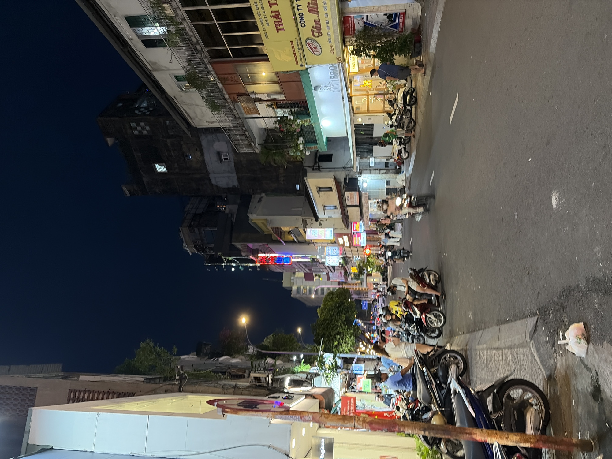 In Thủ Đức, Vietnam, at night, a street scene unfolds with a dark sky overhead. A sidewalk is lined with parked motorcycles and a few pedestrians. On the left, a building with a white facade and yellow accents is visible, featuring a sign with a red and white circular graphic. Further down the street, other buildings rise, displaying various signs and illuminated windows. Red and neon signs are prominently displayed on the buildings, indicating businesses. A street lamp casts a warm glow on the scene. The street itself is paved and appears to be a busy thoroughfare, with more motorcycles and a few individuals walking. One person is sitting on a motorcycle, looking at their phone, while others are standing or walking. The overall impression is a lively urban environment at night, characteristic of a commercial area in Thủ Đức.