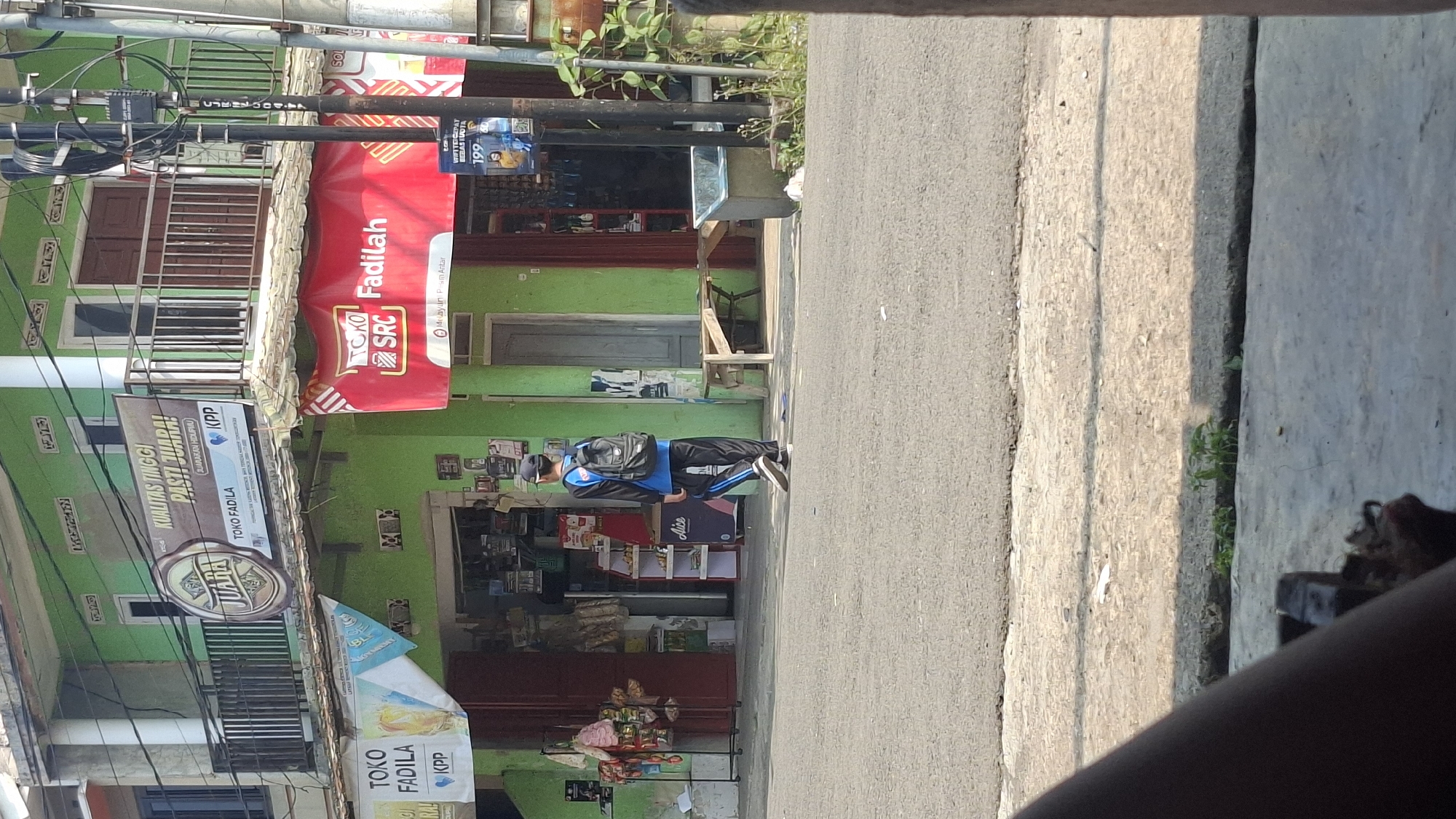 The image depicts a street scene in Cilaku, Indonesia, featuring a green two-story building with commercial establishments on the ground floor. A young male, wearing a blue long-sleeved shirt, black pants with blue stripes, and a black backpack, walks away from the camera on the paved road in front of the building. Above the ground floor shops, a balcony with metal railings is visible. Multiple banners and signs are displayed on the building's facade. To the left, a banner reads "TOKO FADILA KPP". Above this, another banner displays a circular logo with text that appears to include "JUARA". To the right of the "JUARA" banner, text reads "KUALITAS TINGGI PASTI JUARA!". Another prominent red banner on the ground floor proclaims "TOKO SRC Fadilah". A utility pole with multiple wires stands to the right of the building. The environment includes a street, a sidewalk, and some vegetation. The lighting suggests it is daytime.