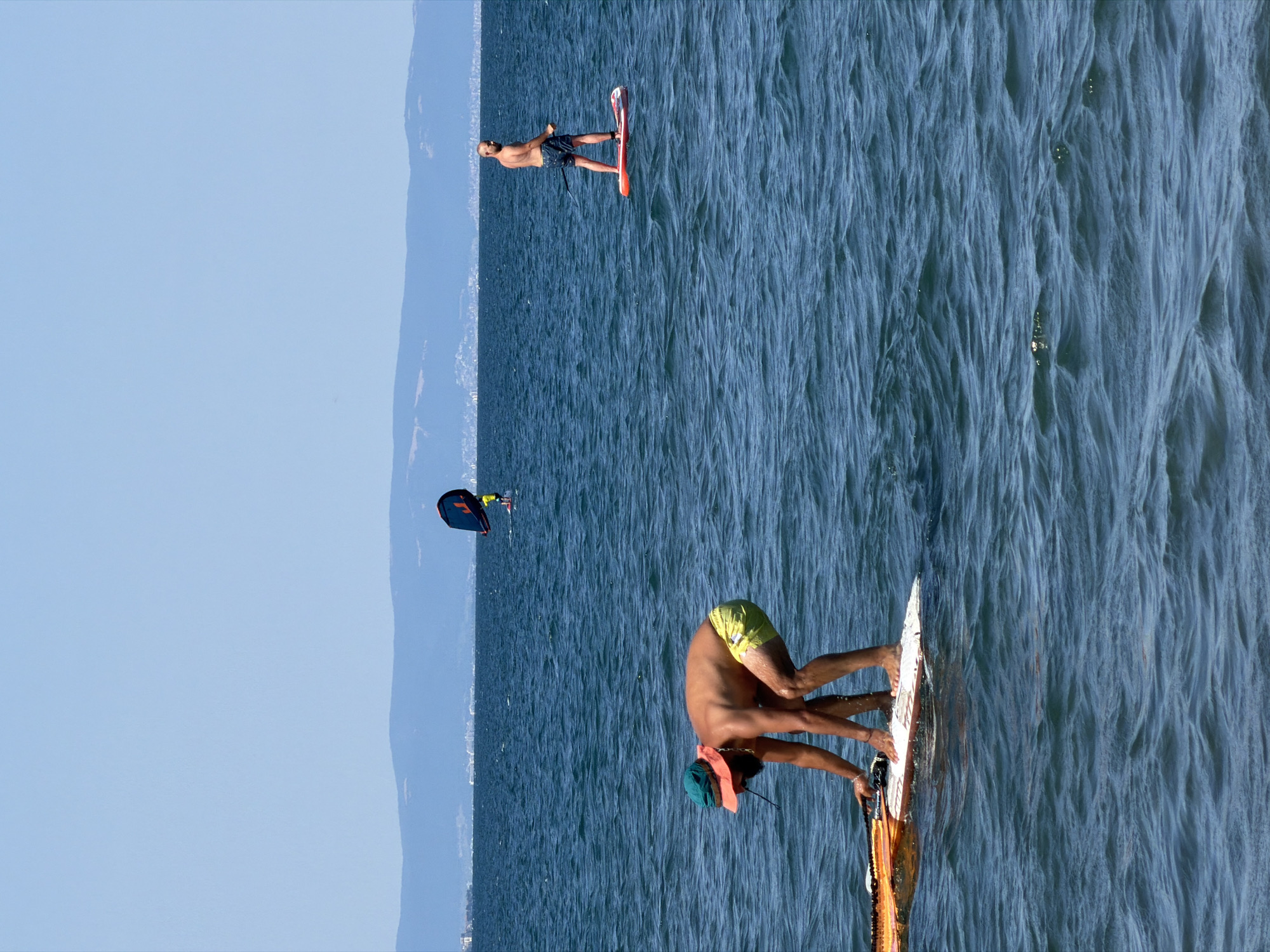 The image captures a sunny day at the sea in Atiya, Bulgaria, with several individuals engaged in water sports. In the foreground, a man wearing a green hat and yellow shorts is crouched on a paddleboard, appearing to adjust equipment. Further out in the water, another man stands on a paddleboard, while a third person is visible in the distance on a paddleboard with a wing sail. The sea is a deep blue, with moderate waves, and a distant coastline with buildings and hills is visible under a clear blue sky.