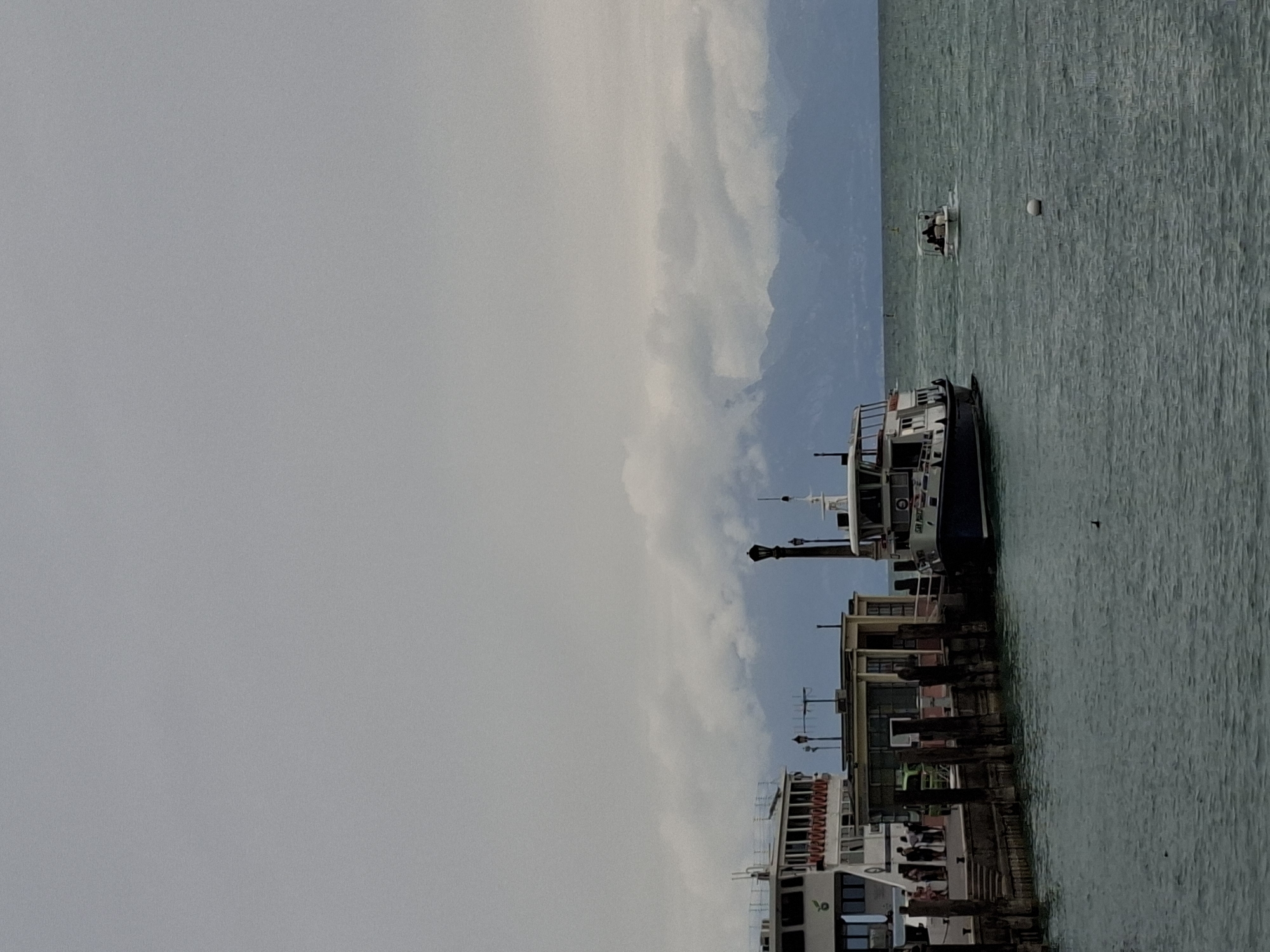 This is a photograph taken at the lakefront in Peschiera del Garda, Italy. In the foreground, the dark green, choppy water of Lake Garda is visible. A white spherical buoy floats in the water on the right side of the frame. Two small dark birds are also visible in the water.
On the left side of the image, a portion of a large white ferryboat is docked at a pier. The ferry has multiple decks and many windows. People can be seen on the pier and on the ferry. The pier is made of wood and concrete, with vertical wooden pilings supporting it. Adjacent to the ferry and pier is a small, light-colored building with large windows.
To the right of the pier and building, a smaller dark-hulled boat with a white upper structure is also docked. This boat appears to be a passenger ferry, with the name "SAN MARCO" visible on its side.
Further out in the water, a small white motorboat with three occupants is moving away from the shore.
In the background, the hazy outlines of mountains are visible against a cloudy, overcast sky. The clouds appear to be low-lying, partially obscuring the mountain peaks. The overall lighting suggests a cloudy or overcast day.